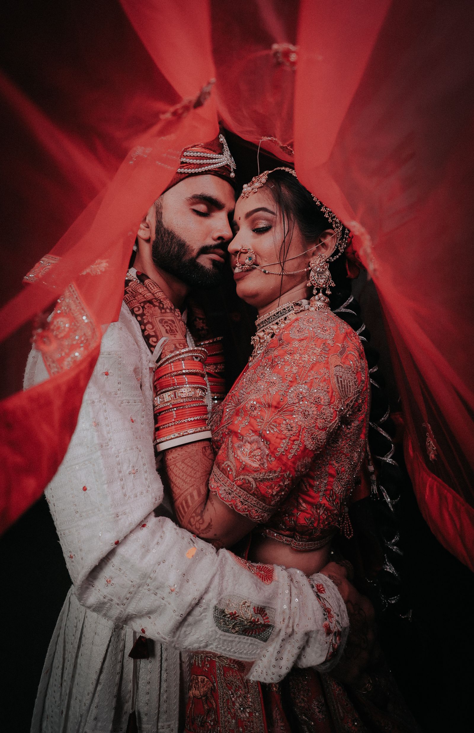 A couple embracing beneath a backlit red dupatta held up over both of them.