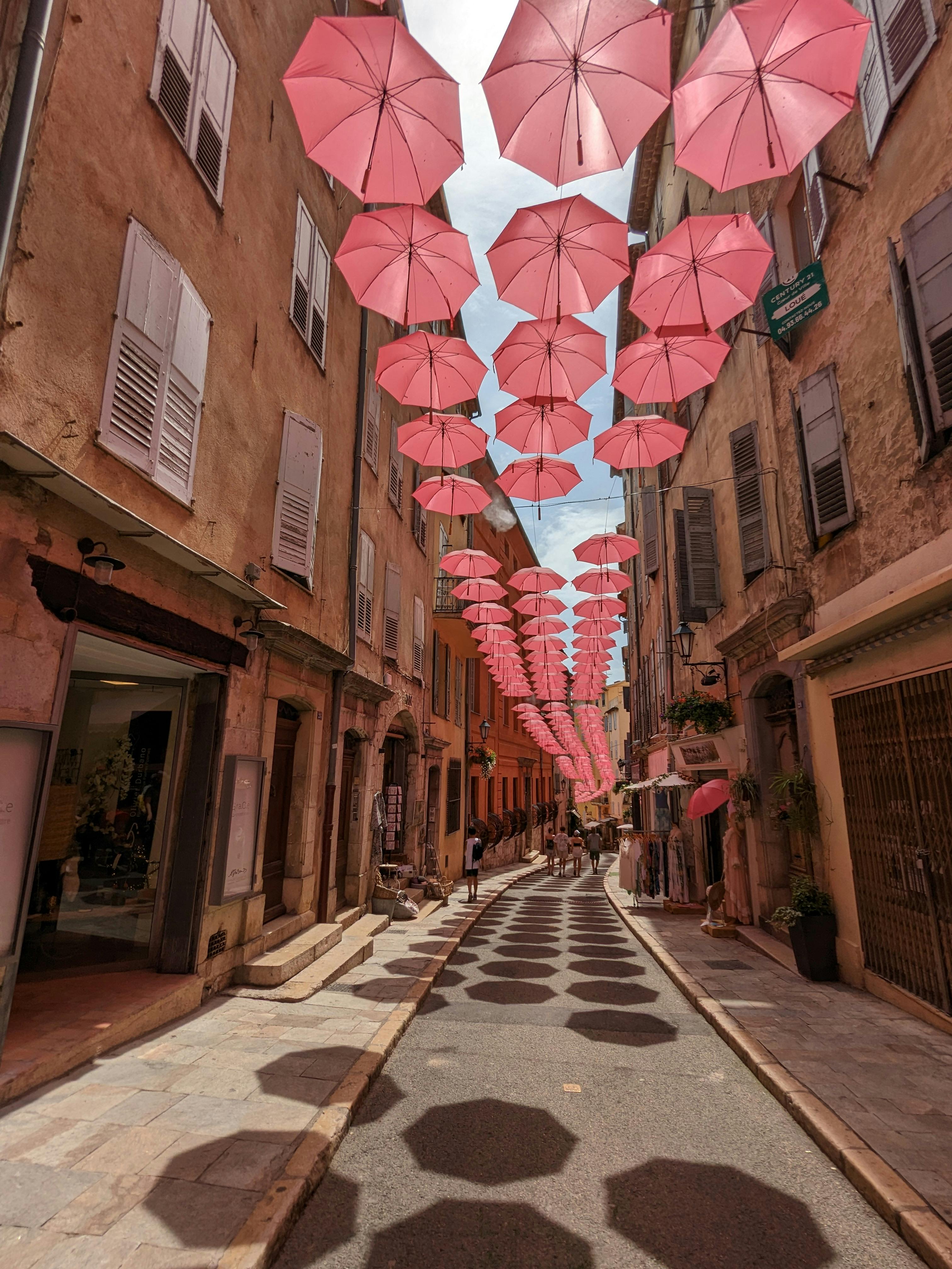 A charming street in Grasse, France adorned with a pink umbrella canopy creating whimsical shadows.