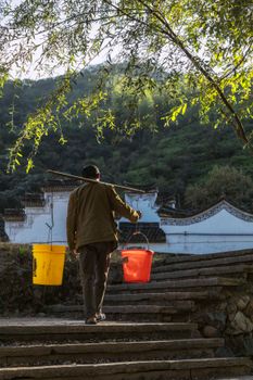 Back view faceless male in worn out clothes carrying plastic buckets with clean water by using shoulder pole while walking up on shabby street stairs in countryside