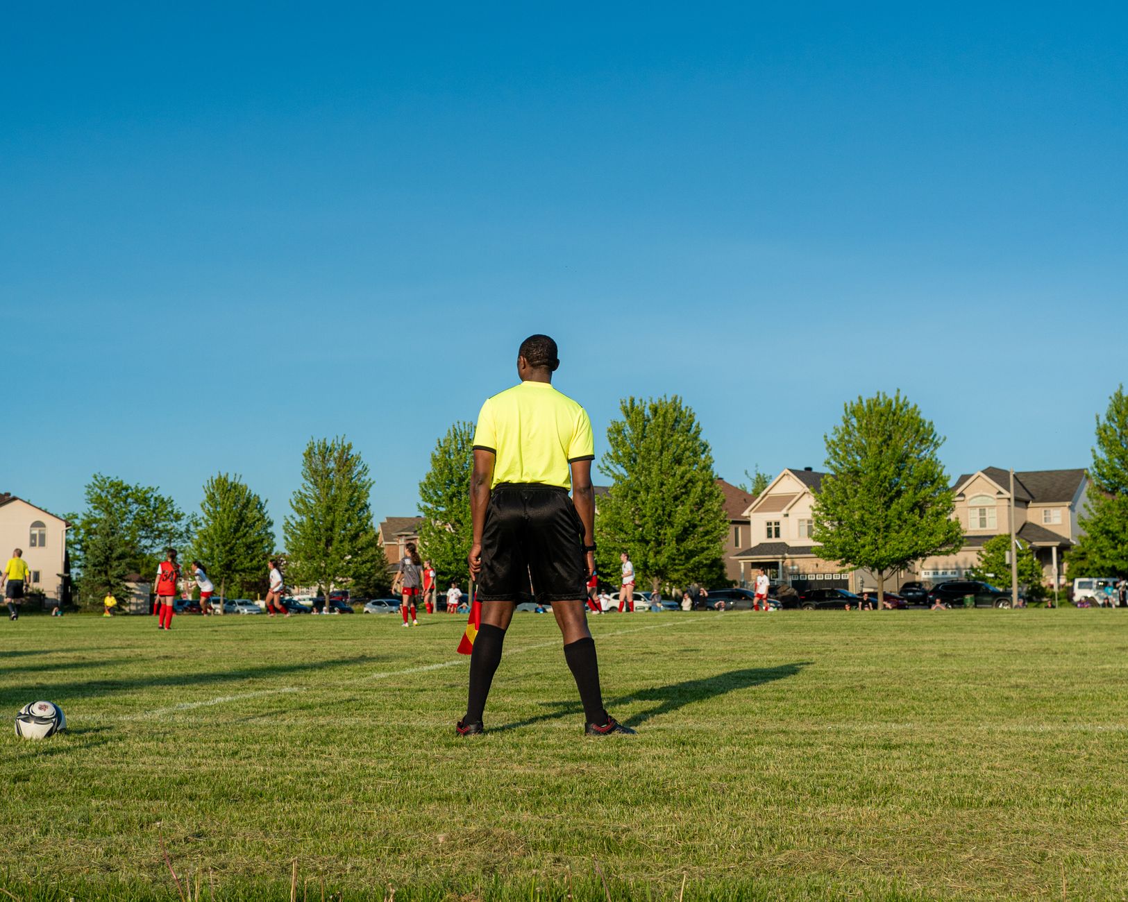 Referee oversees a soccer match on a sunny day in a suburban neighborhood.