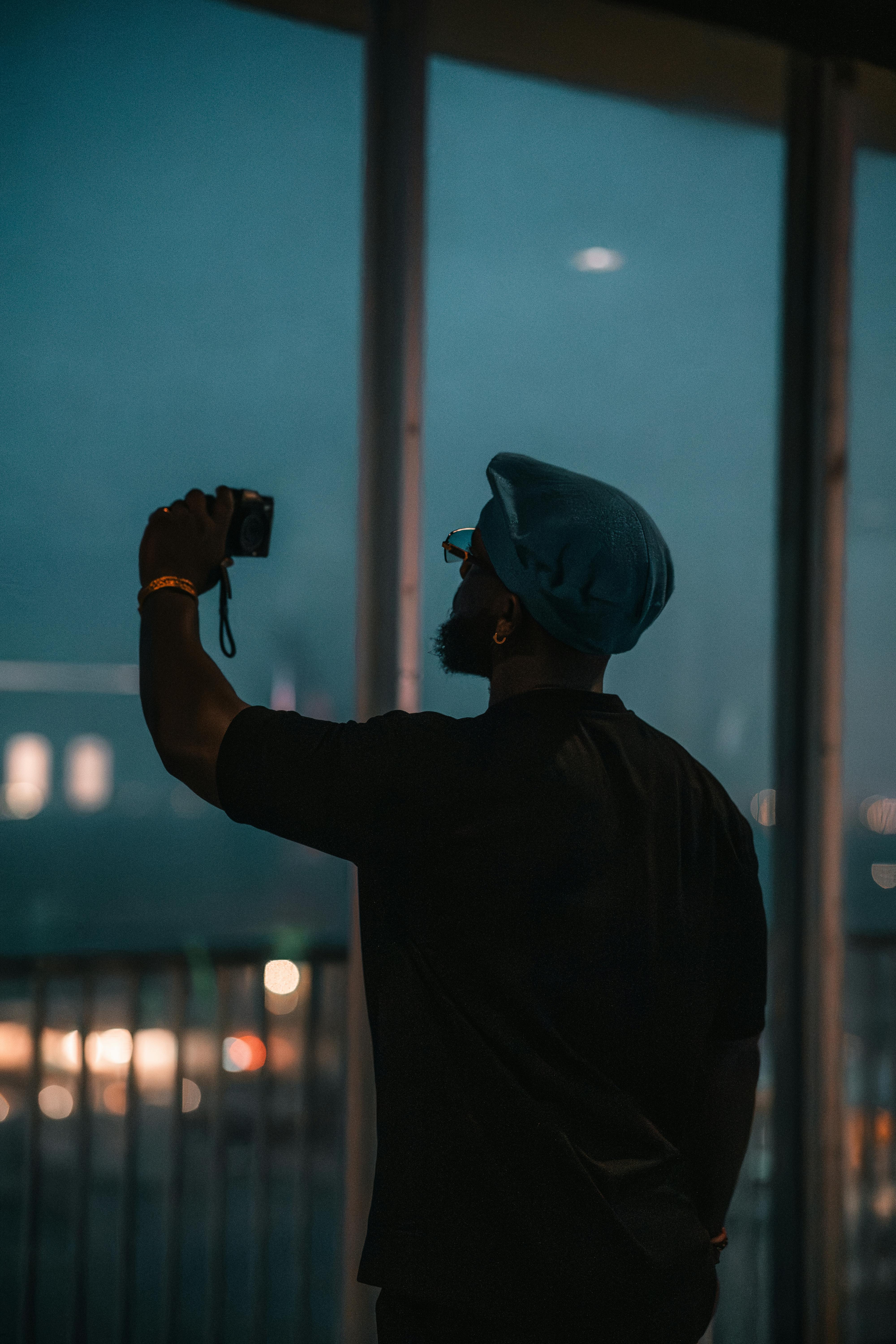 Kostenlos Silhouette eines Fotografen, der abends durch ein Fenster eine städtische Landschaft einfängt. Stock-Foto