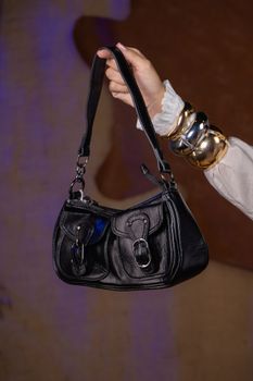 A fashionable black leather handbag held by a hand wearing a stylish bracelet against a soft focus background.
