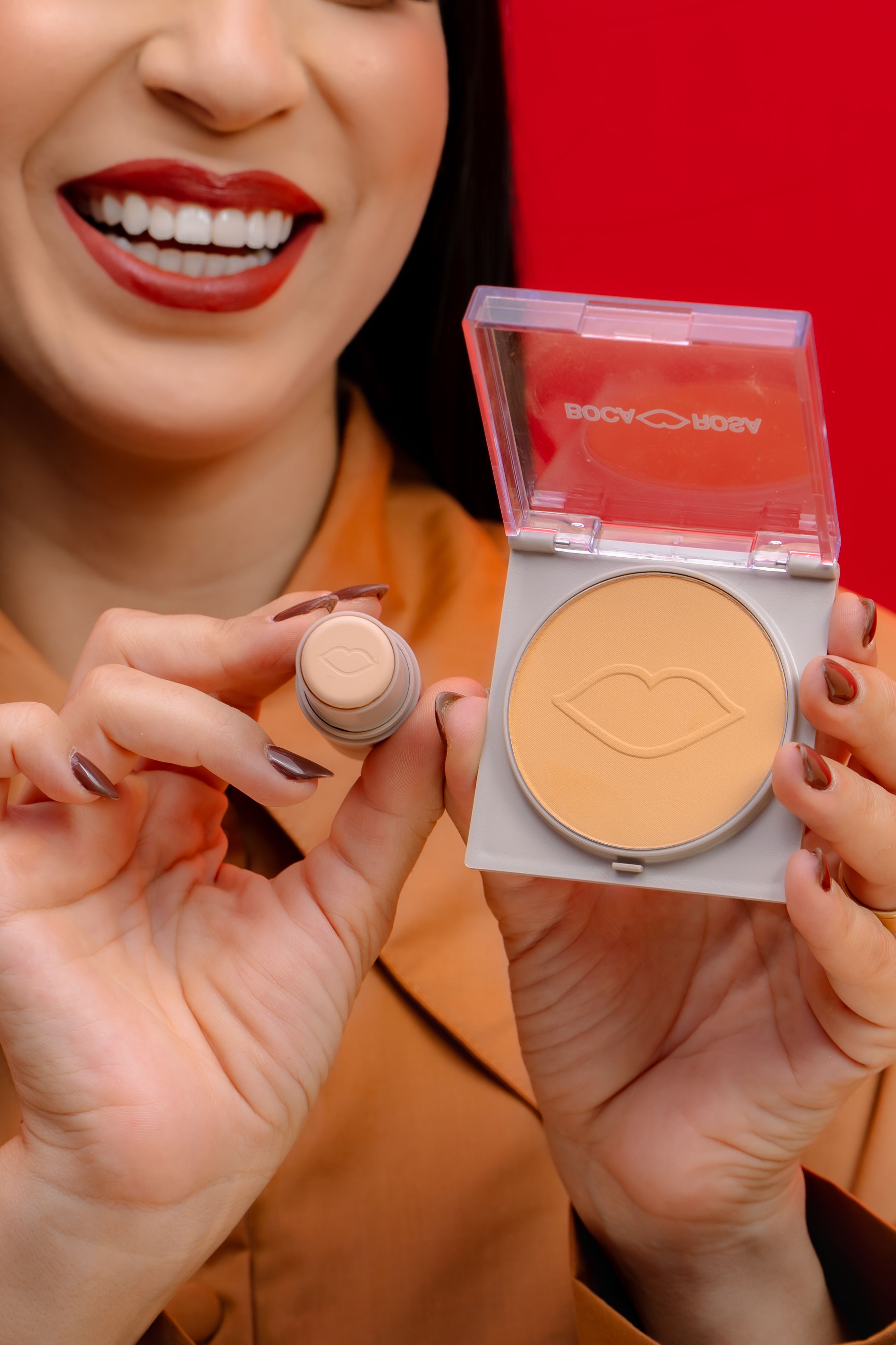 Closeup of a woman showcasing makeup products with a smile against a red background.