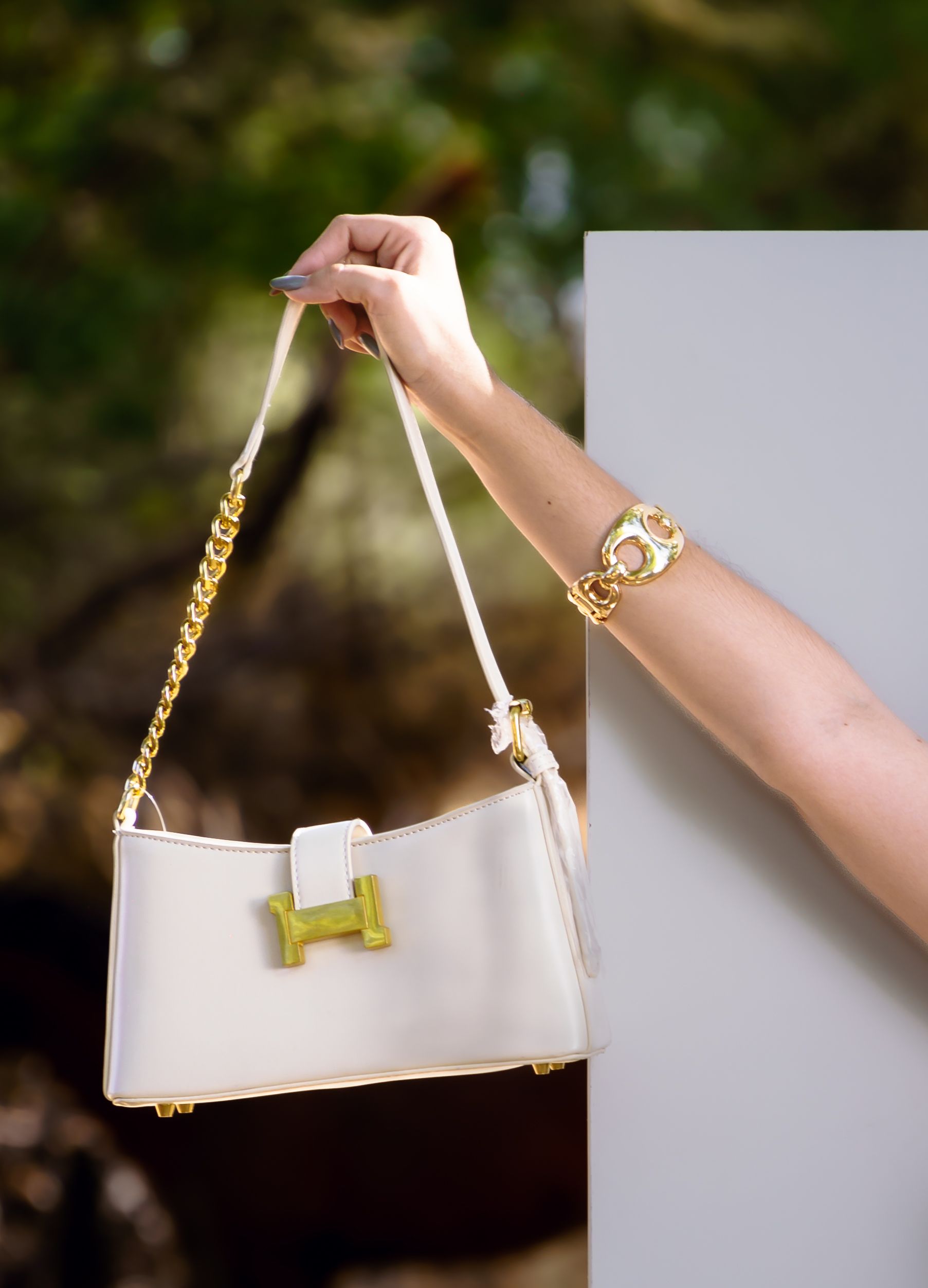 Uma elegante bolsa branca e dourada segurada pela mão de uma mulher usando uma pulseira de ouro, ao ar livre.