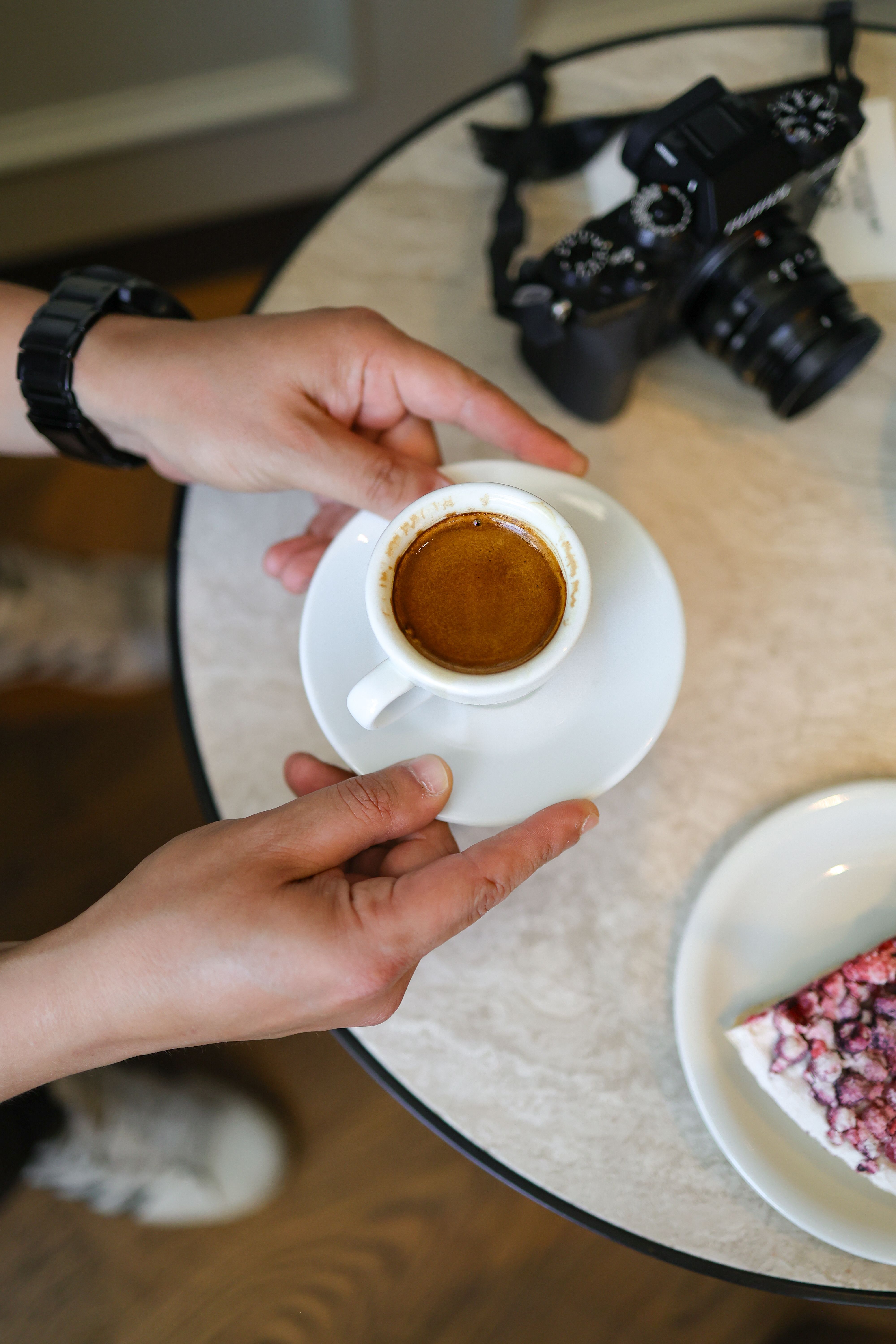 Close-up of hands holding an espresso cup on a café table with a camera and dessert.