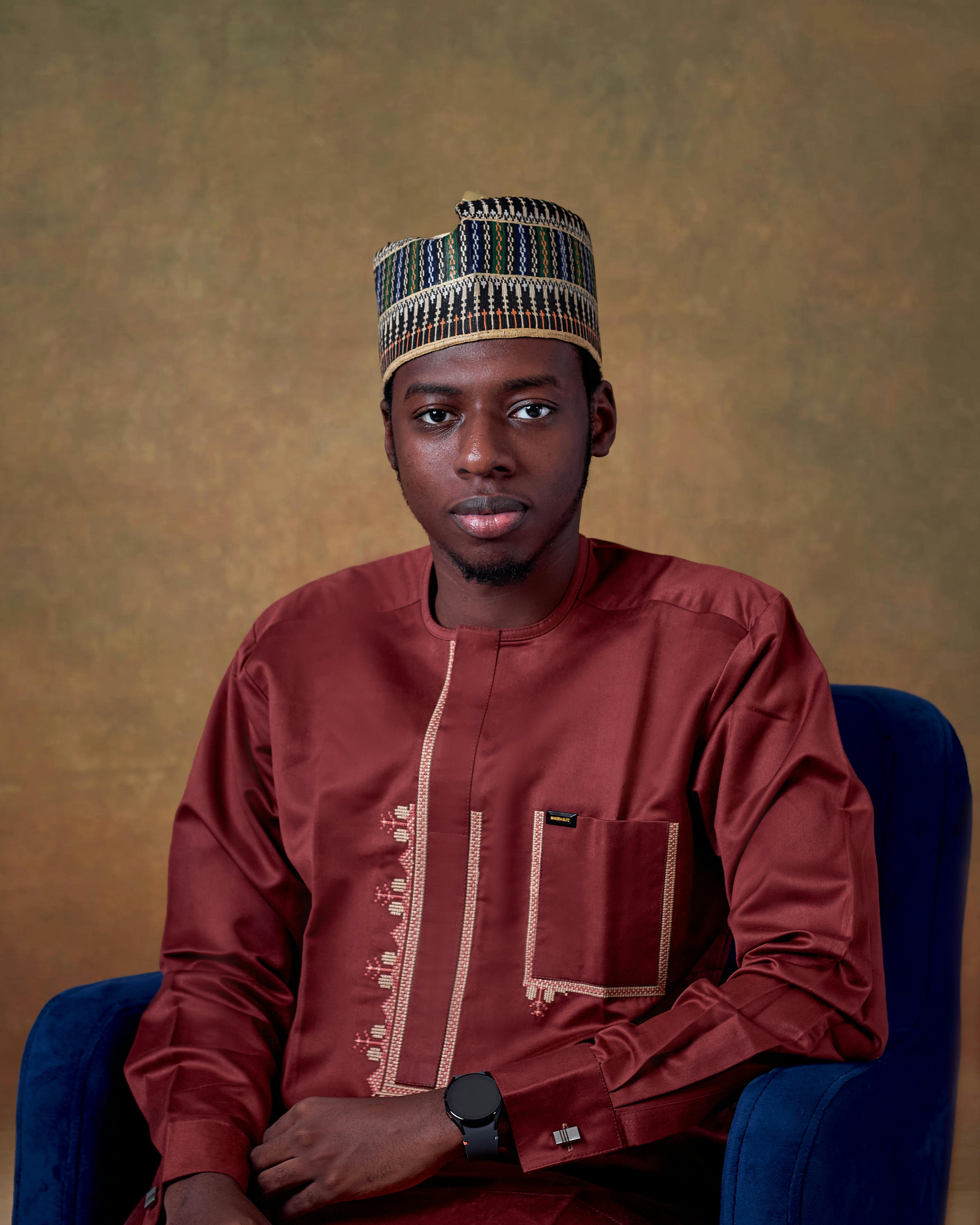 Man wearing a traditional Nigerian outfit with a patterned hat, seated in a studio setting.