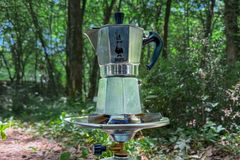 Moka pot brewing coffee on a portable stove amidst forest greenery, capturing a serene outdoor experience.