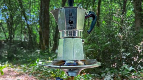 Moka pot brewing coffee on a portable stove amidst forest greenery, capturing a serene outdoor experience.