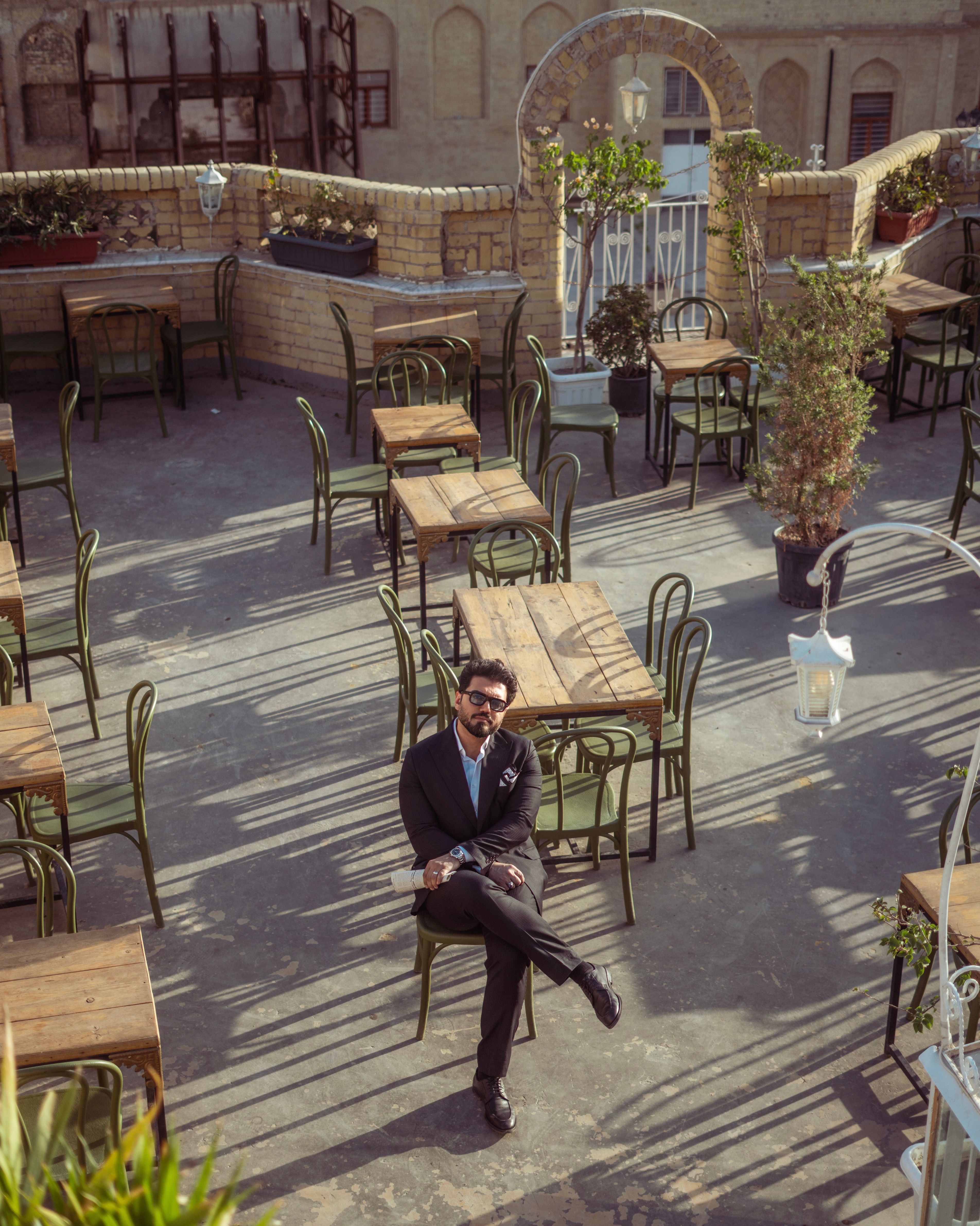 A stylish man in a suit sits alone in an outdoor cafe in Baghdad, Iraq, surrounded by empty tables and chairs.