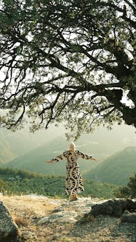 A woman in a patterned dress stands under a tree, arms outstretched, overlooking mountains.