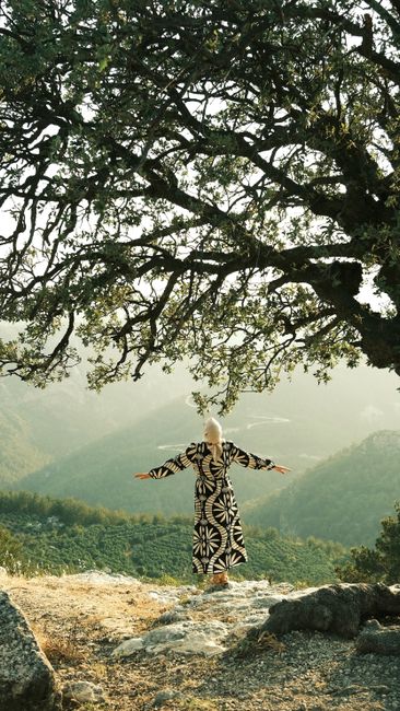 A woman in a patterned dress stands under a tree, arms outstretched, overlooking mountains.