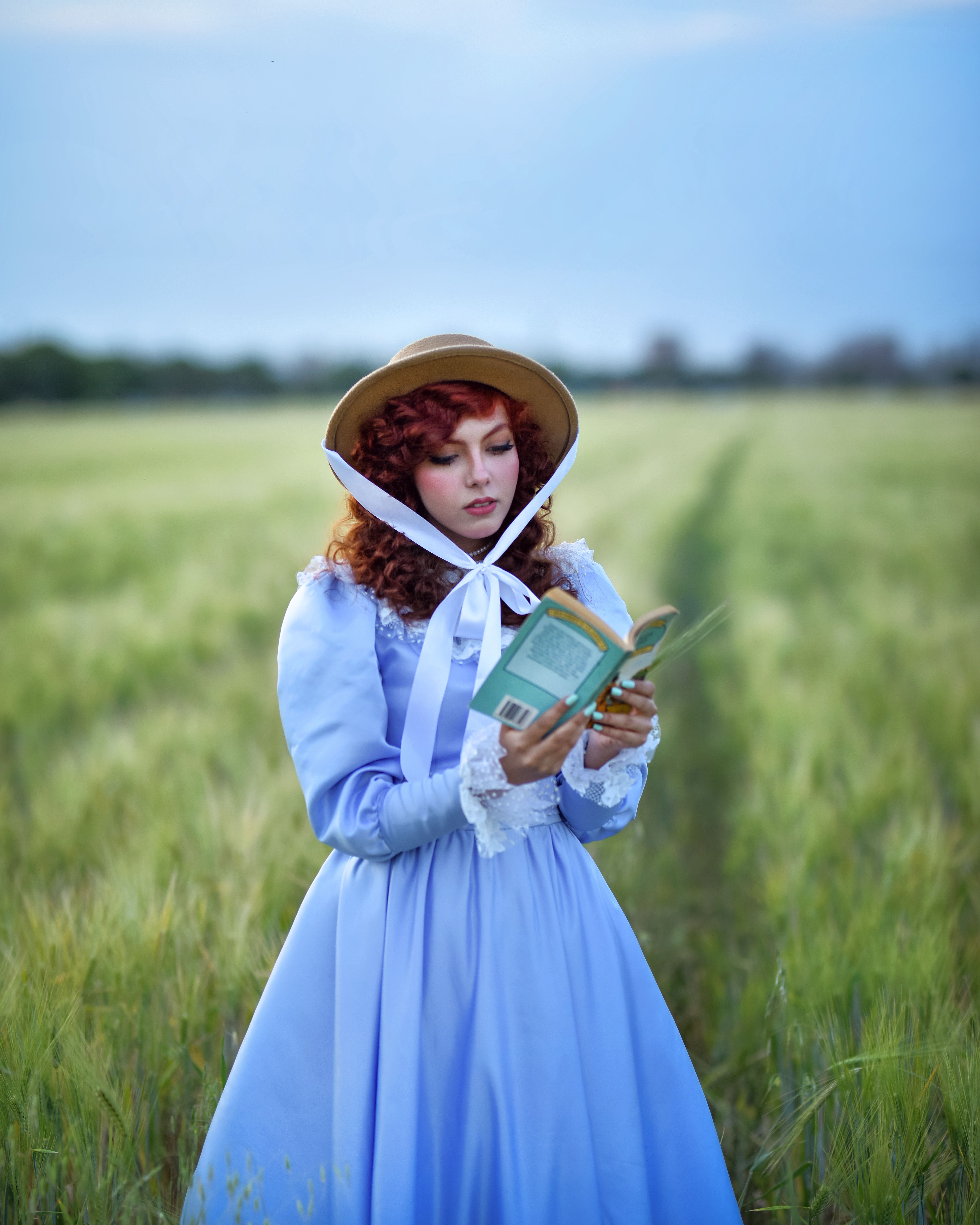 A woman in a vintage blue dress reads a book in a green field under a clear sky.