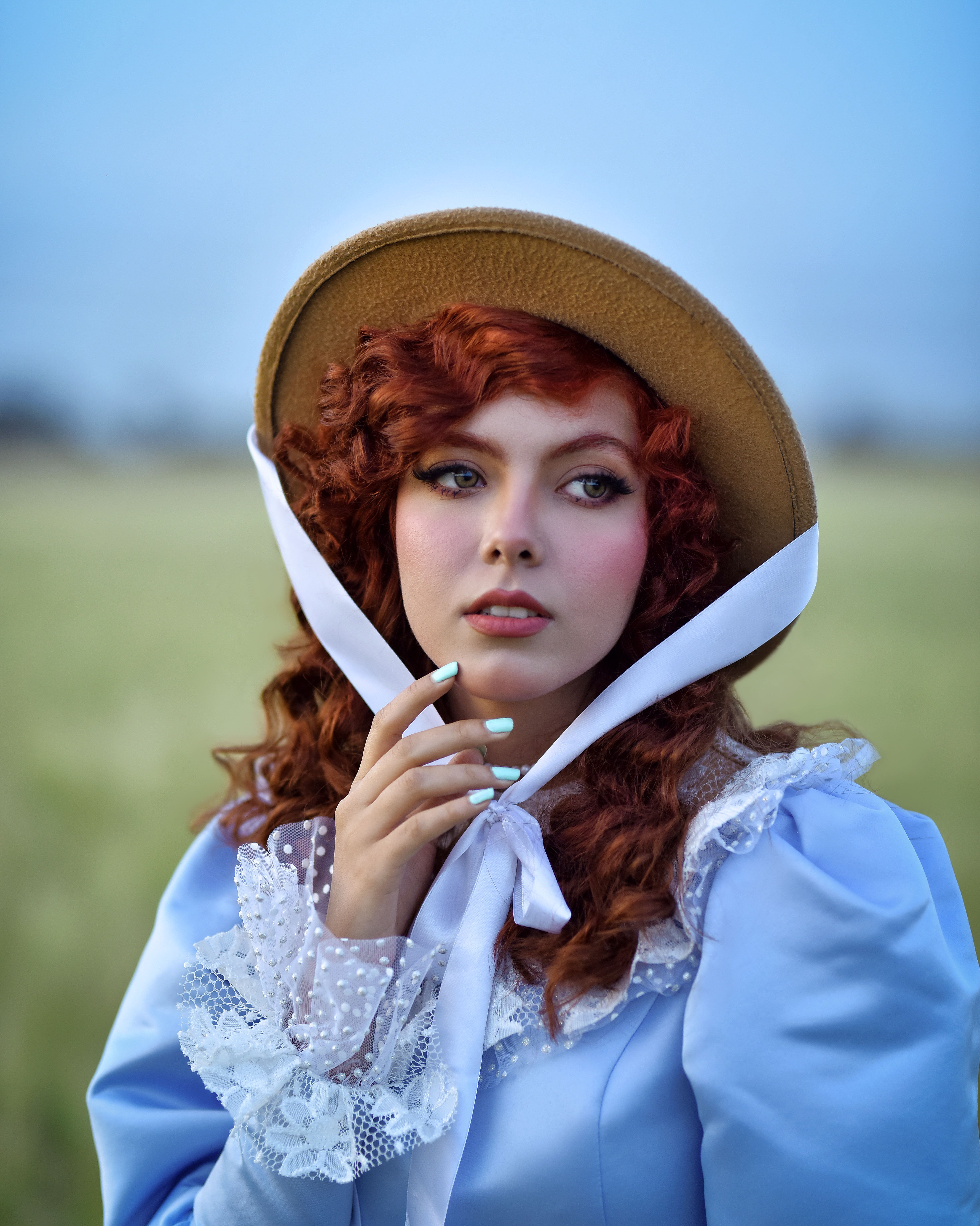 Portrait of a young woman in vintage fashion with curly red hair wearing a hat in a field.