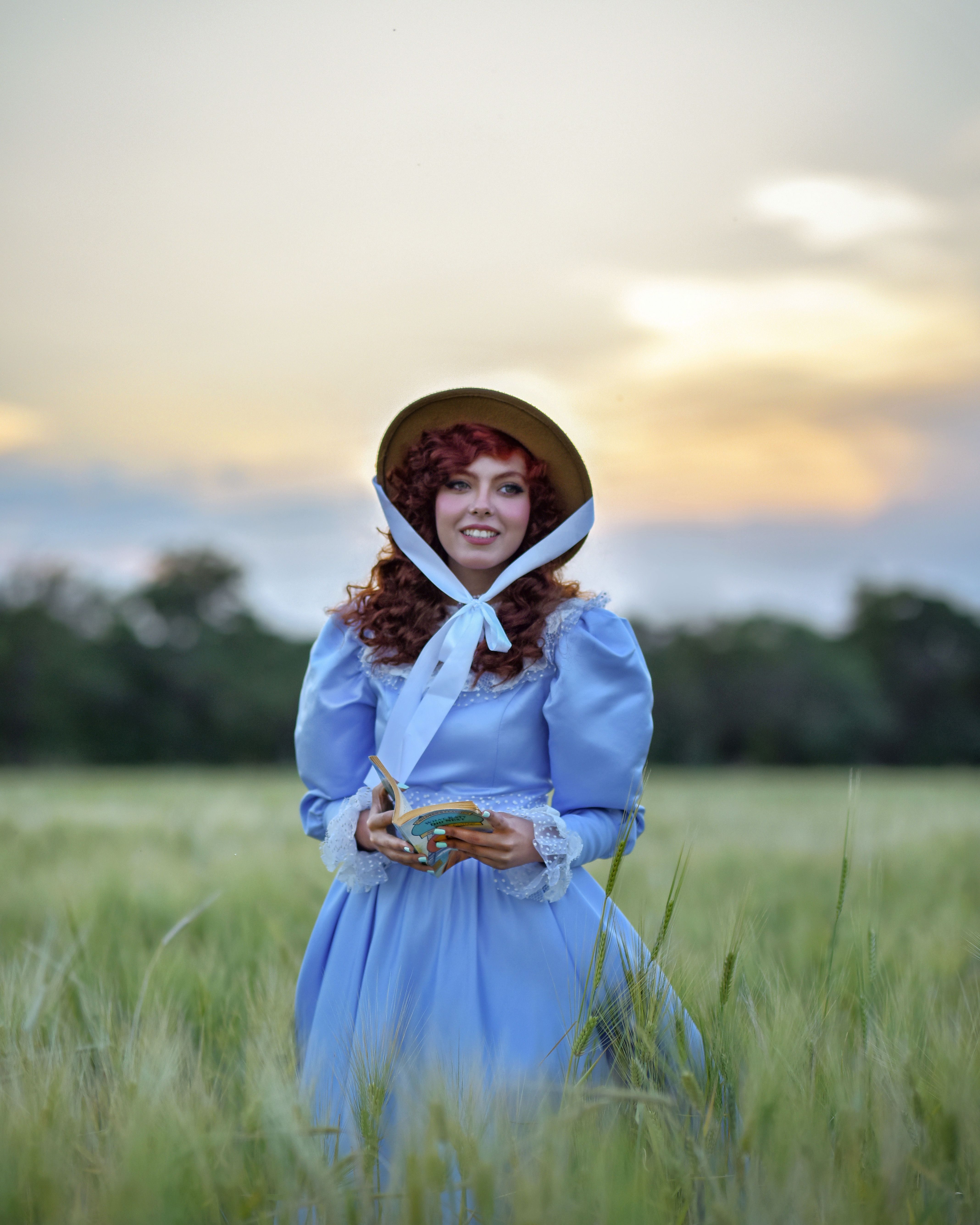 Woman dressed in vintage costume in a field at sunset holding a book, creating a whimsical scene.