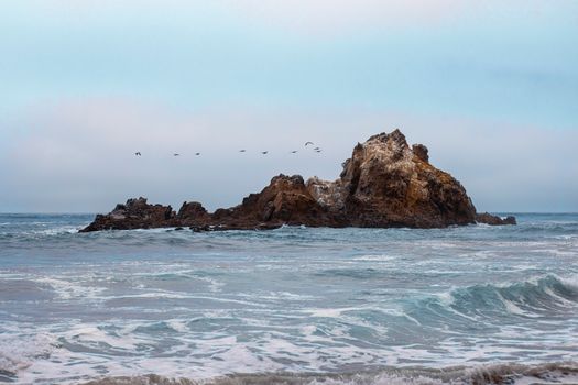 A serene view of a rocky island off the California coast with gentle waves and a flock of birds.