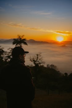 A person gazing at a stunning sunrise over misty mountains and palm trees.