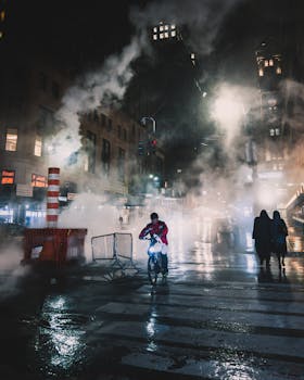 夜晚，一名骑自行车的人穿行在潮湿的城市街道上，四周蒸汽弥漫，城市灯光闪烁