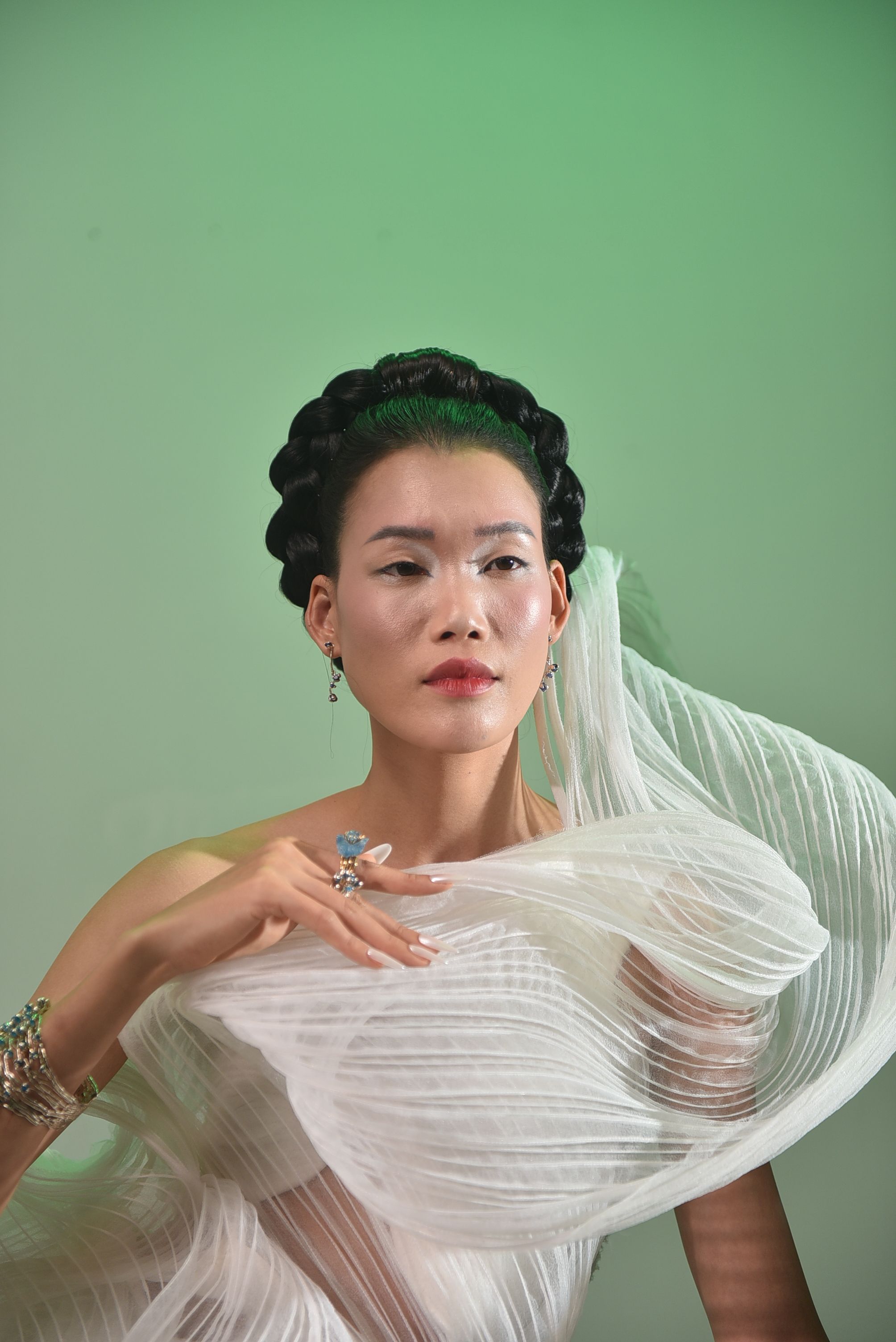 A woman with elegant makeup and hairstyle in a sheer, flowing outfit against a green background.