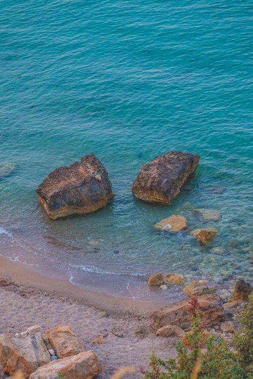 Free A tranquil view of a rocky coastline with crystal-clear blue waters and scattered boulders. Stock Photo