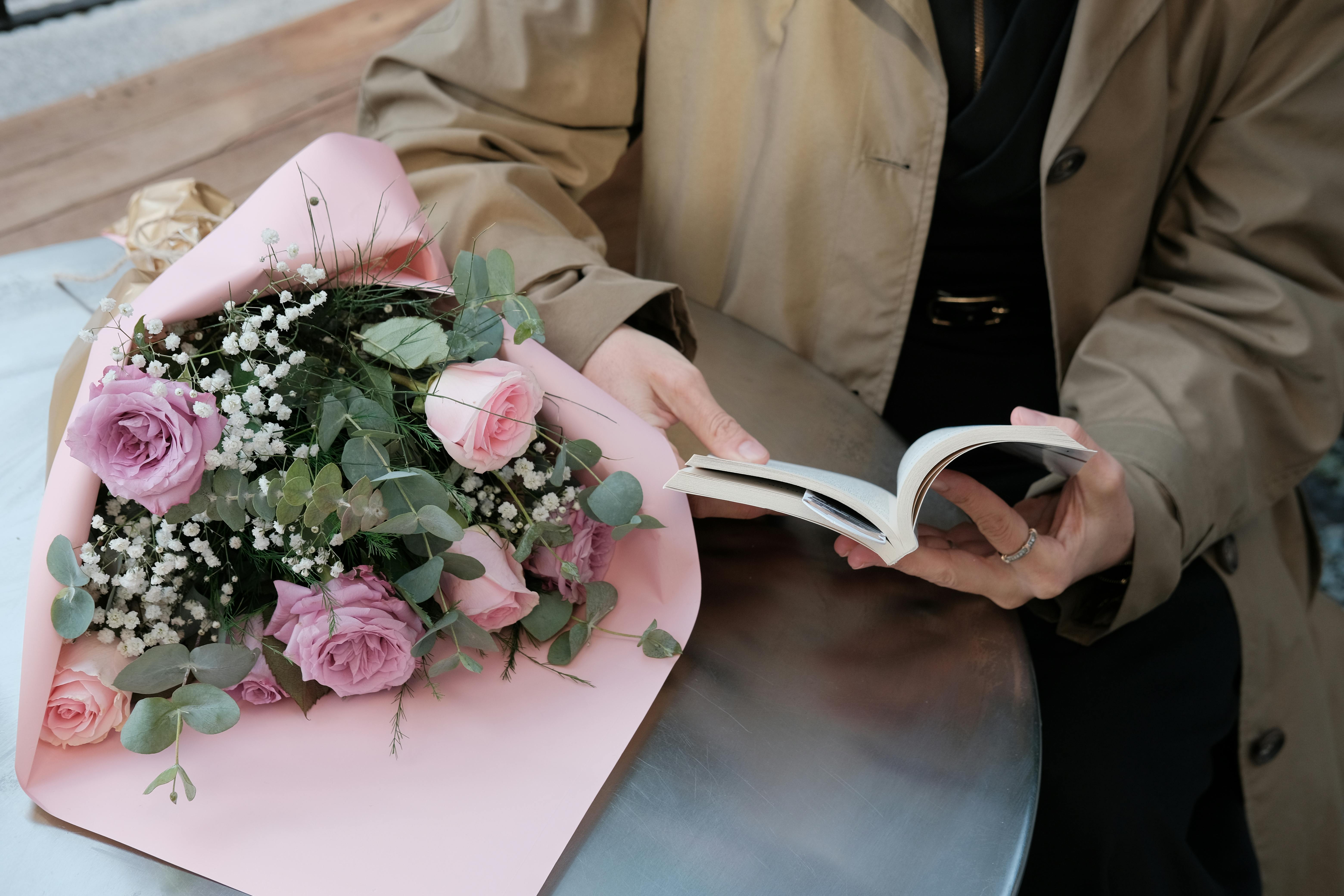A stylish woman in a beige coat reads a book next to a bouquet of pink roses on a cafe table.