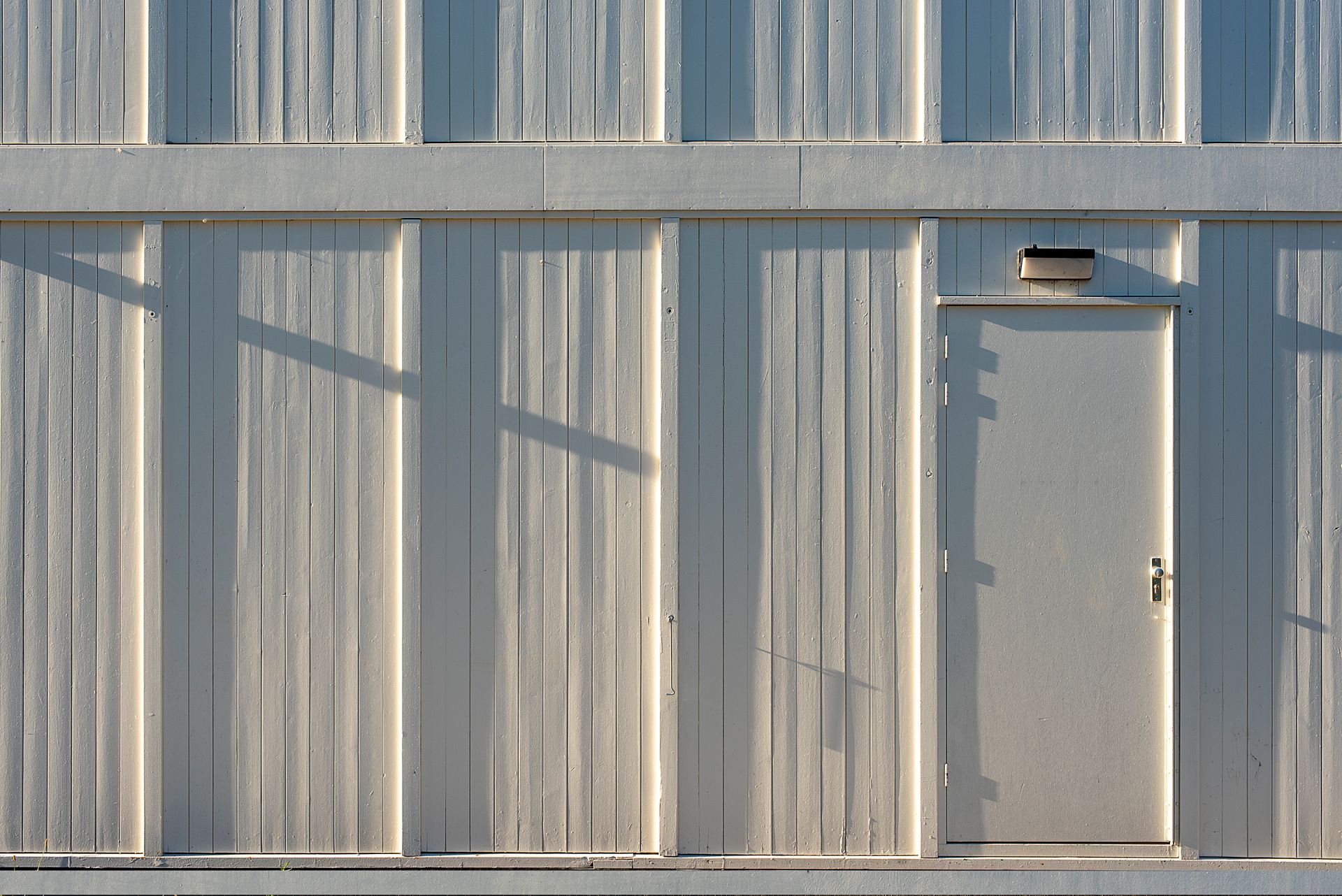 A vertical metal panel wall with a closed door casting geometric shadows in sunlight.