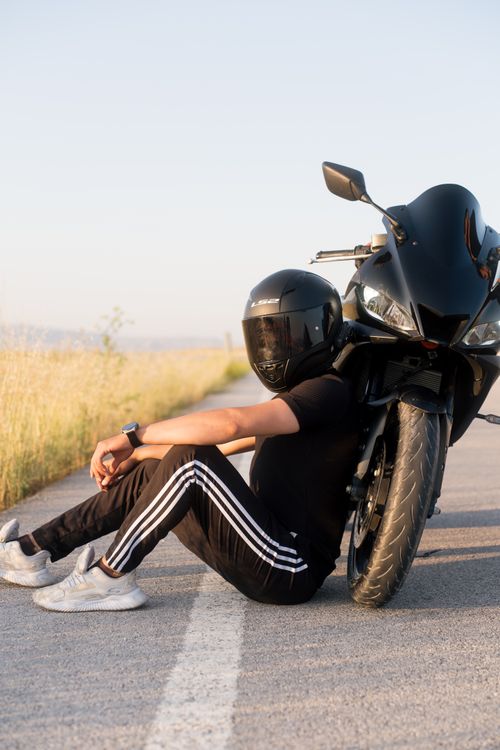 Free Motorcyclist in black helmet sitting by a sleek black sports bike on an open road with a sunny backdrop. Stock Photo