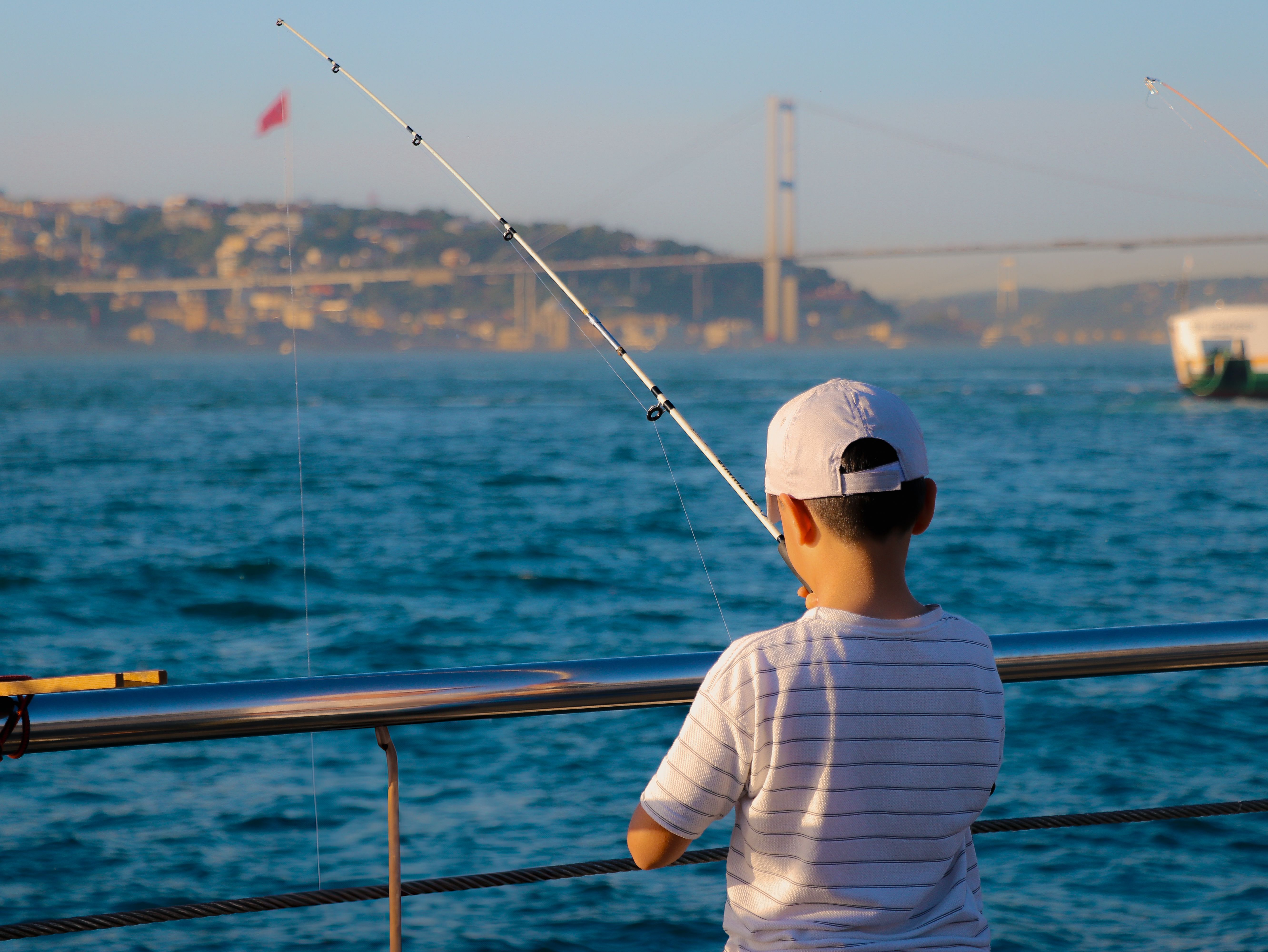 A young boy fishing along the Bosphorus with Istanbul's iconic bridge in the background.