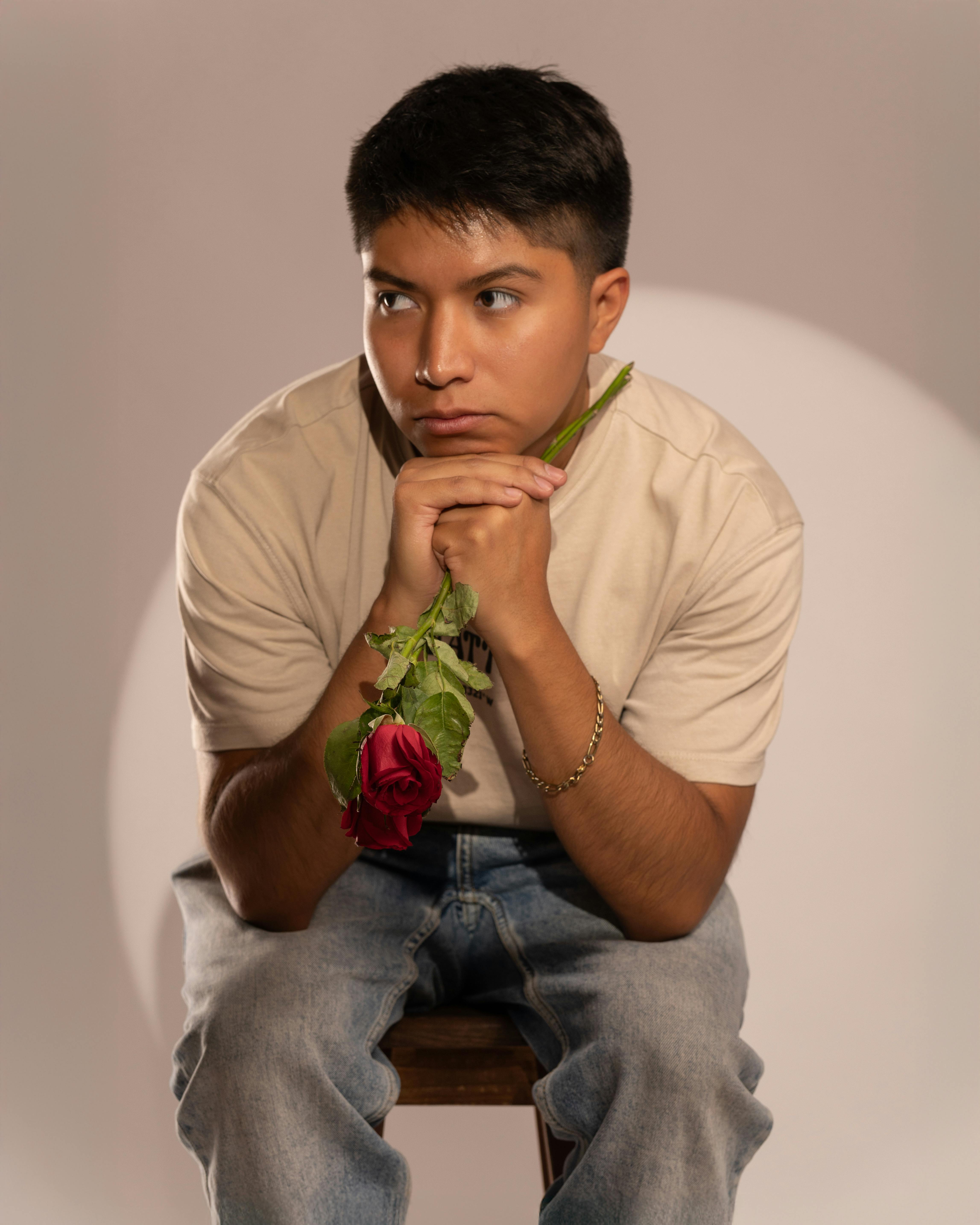 Young man sitting thoughtfully with a red rose in a studio environment, evoking a sense of contemplation.