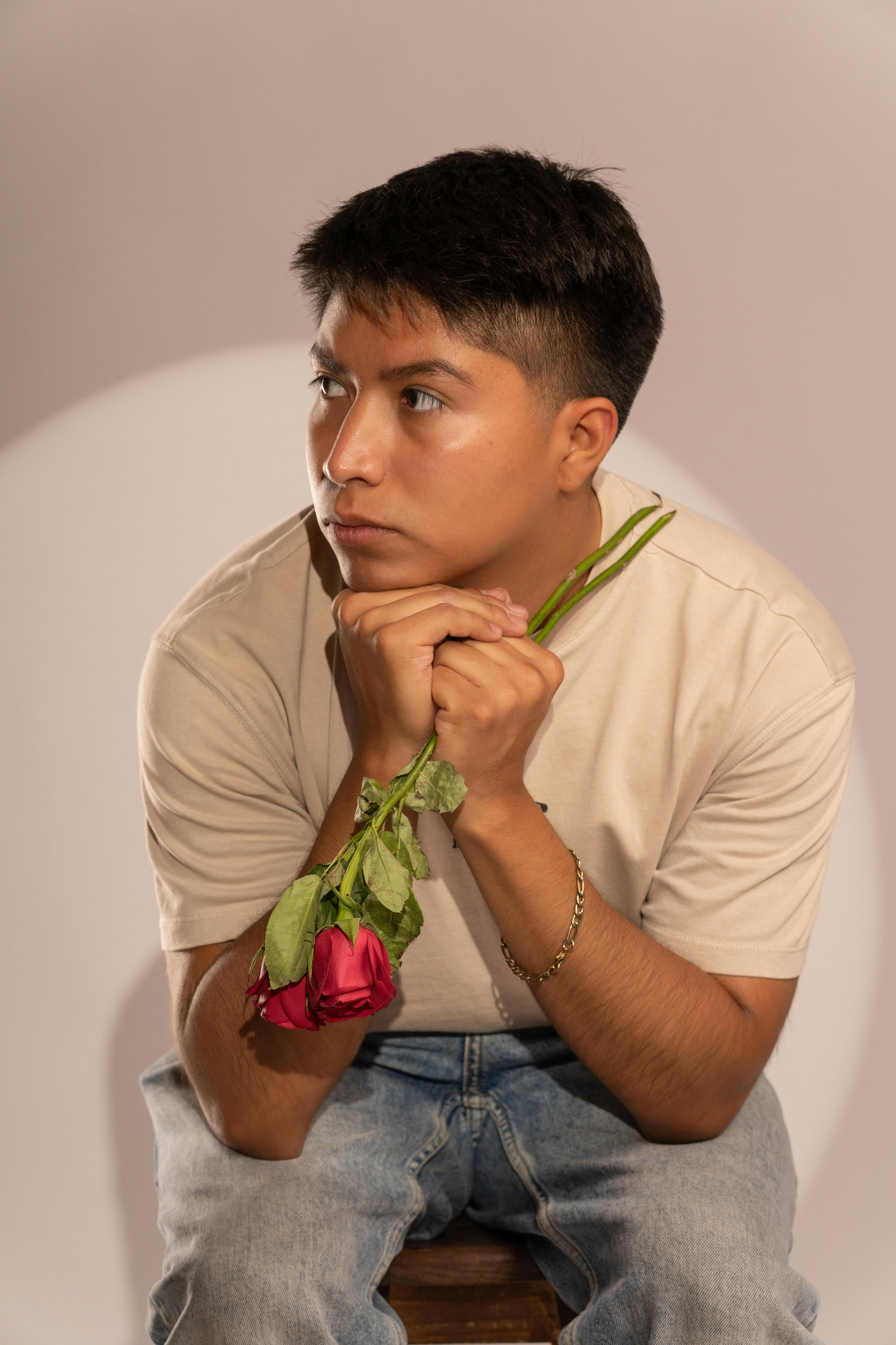 無料 屋内で座り、思索と感情を表現している、赤いバラを持った若い男性の肖像画。 写真素材