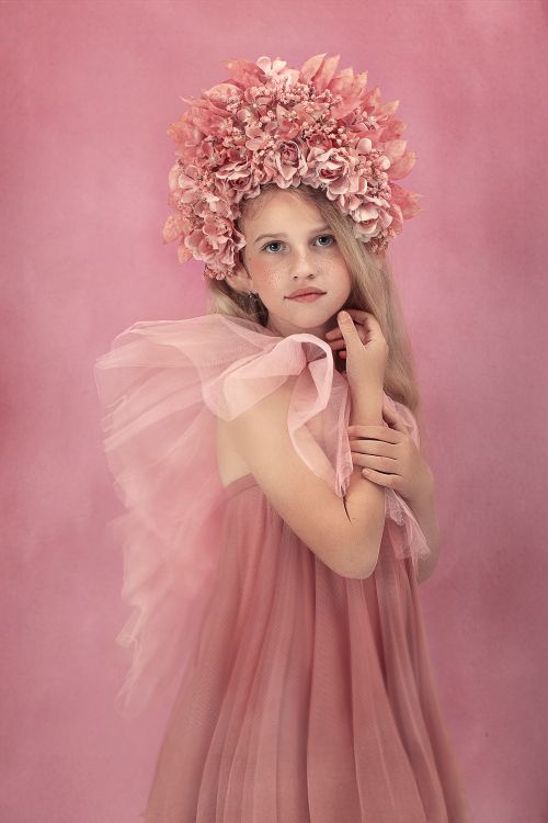 Free A young girl in a pink dress and floral headpiece stands against a pink background, embodying elegance and grace. Stock Photo