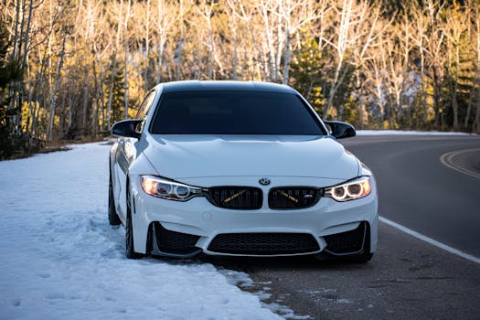 A modern white car parked on a snowy road surrounded by winter trees, front view.