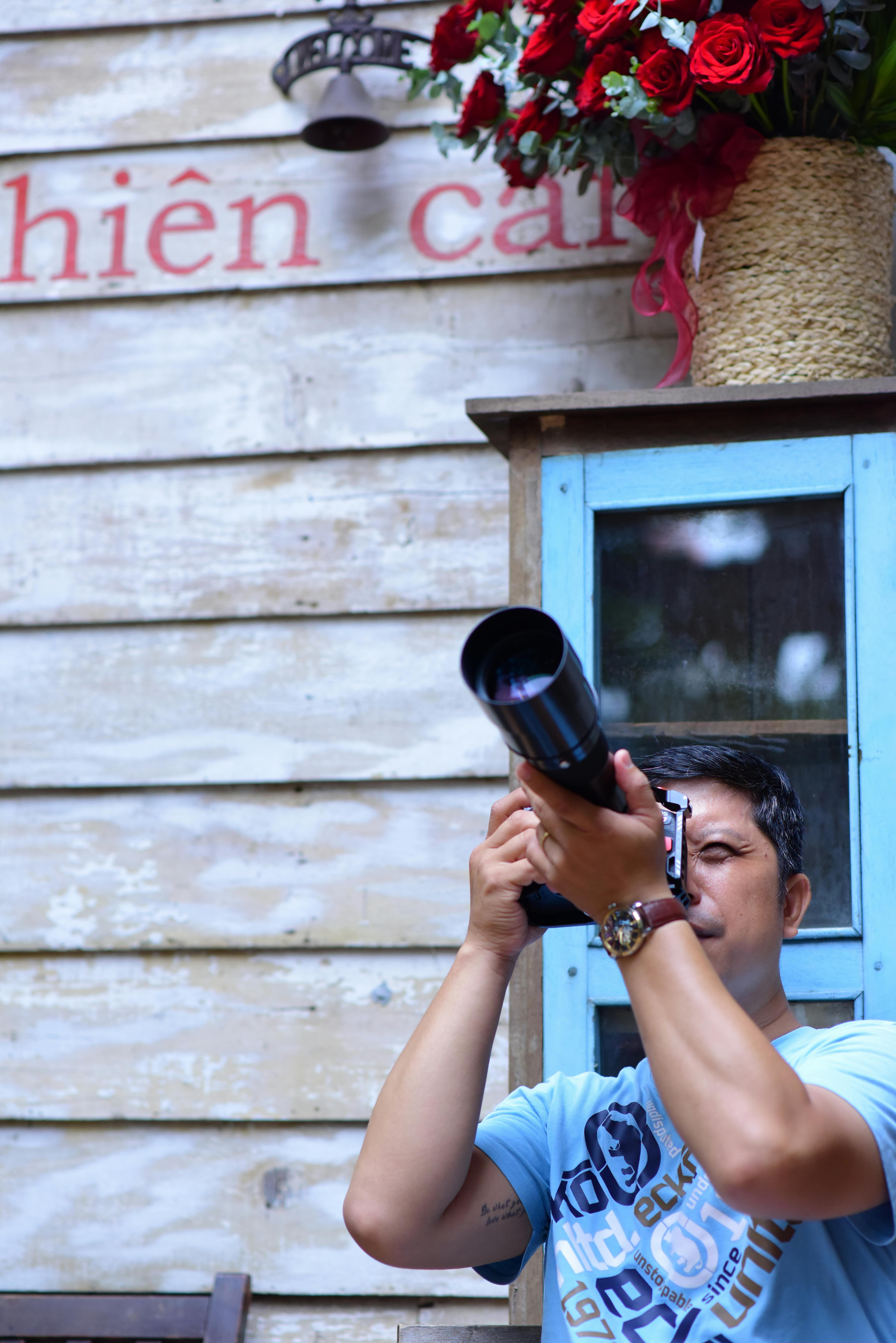 A man aiming a DSLR camera, capturing a scene against a wooden wall and floral decor.