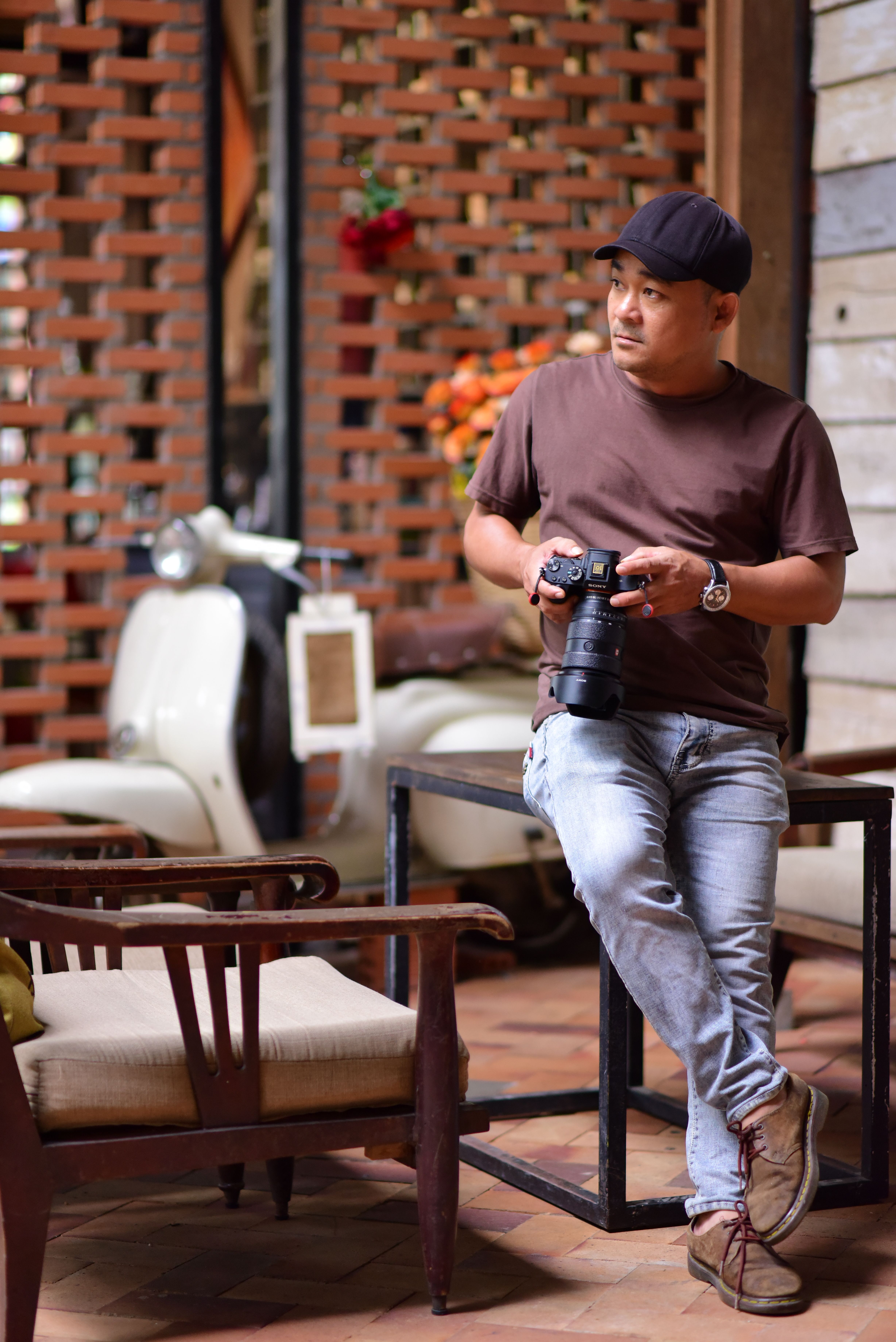 Casual portrait of a man with vintage camera indoors, featuring retro scooter and rustic decor.
