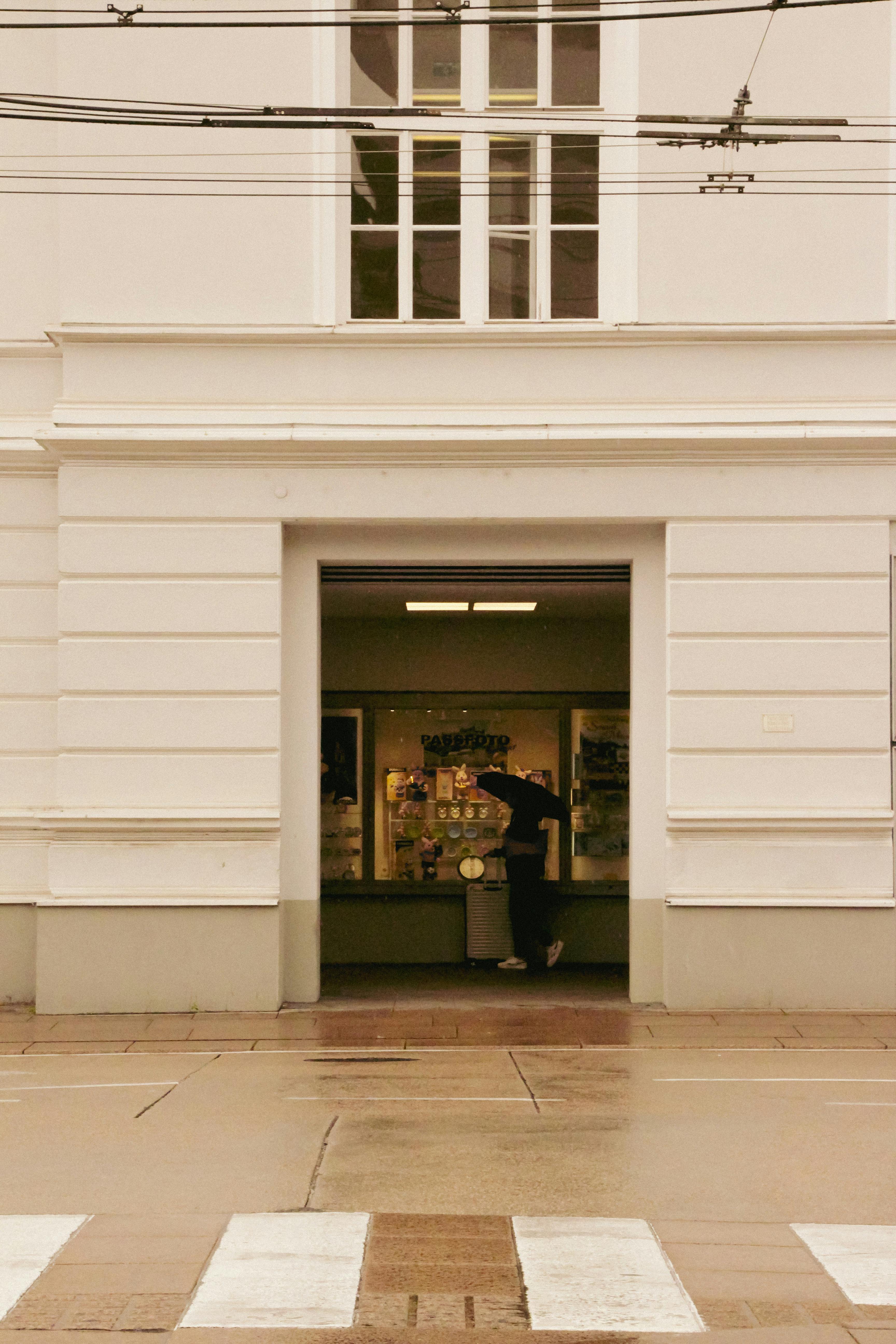 Gratis En person med en paraply går forbi en butiksfacade på en regnvejrsdag i byen. Lagerfoto