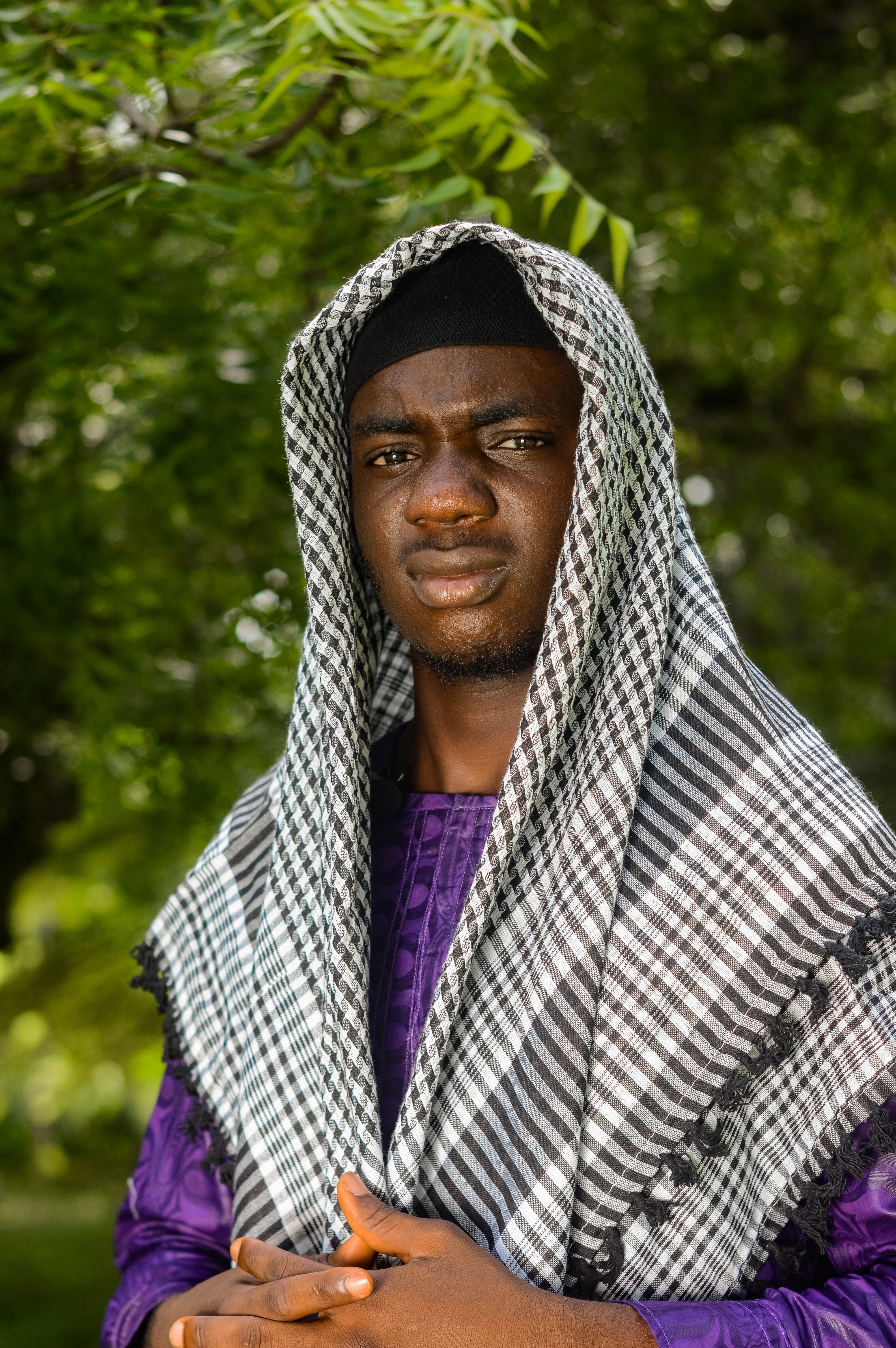 A person in traditional attire with a checkered scarf stands outdoors, surrounded by greenery.