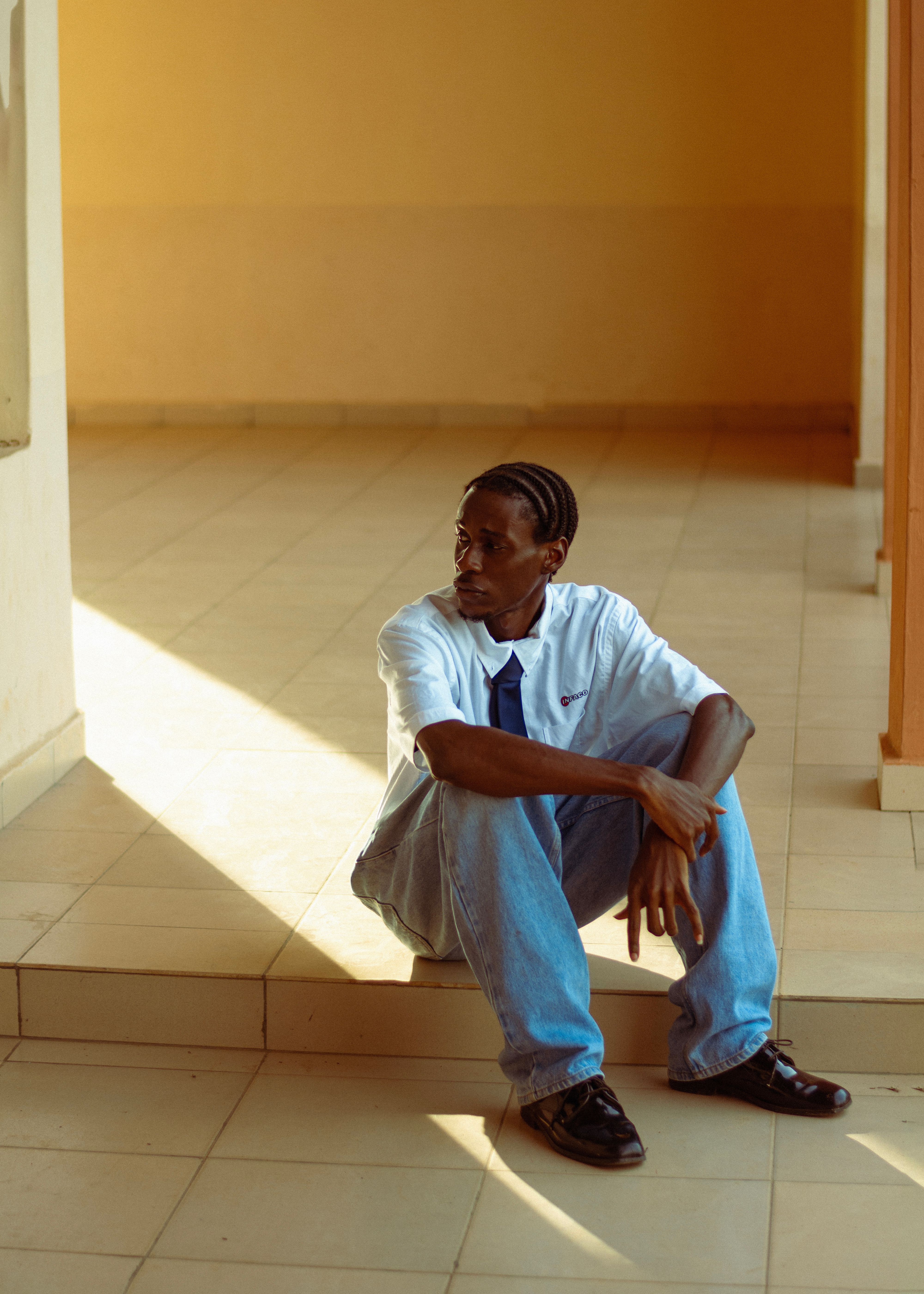 Casual portrait of a young man sitting in a sunlit indoor hallway wearing a white shirt.