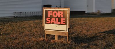 A 'For Sale' sign stands in the grassy yard of an existing home in the late afternoon light.
