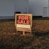 A 'For Sale' sign stands in the grassy yard of an existing home in the late afternoon light.
