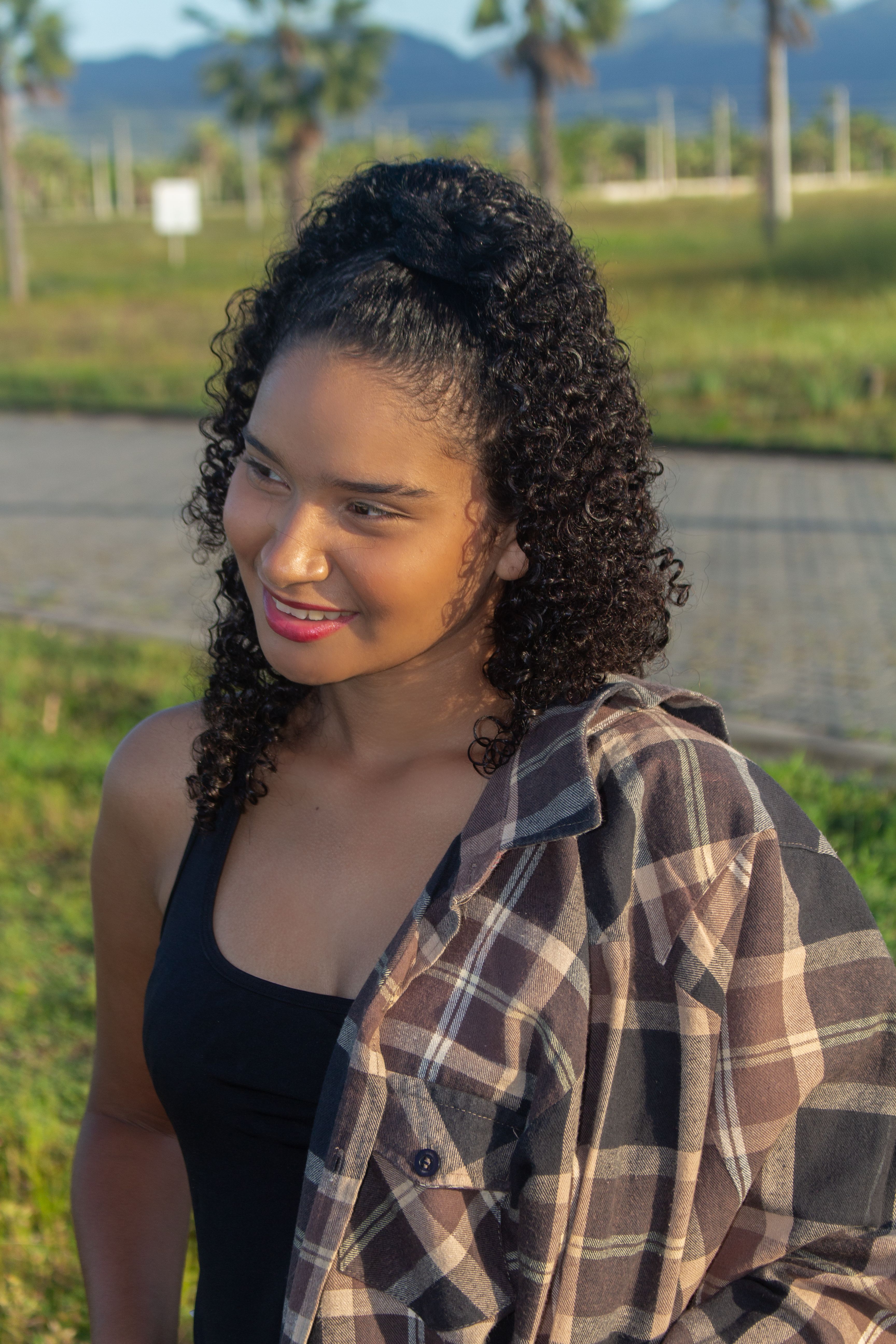 A woman with curly hair smiling outdoors wearing a plaid shirt.