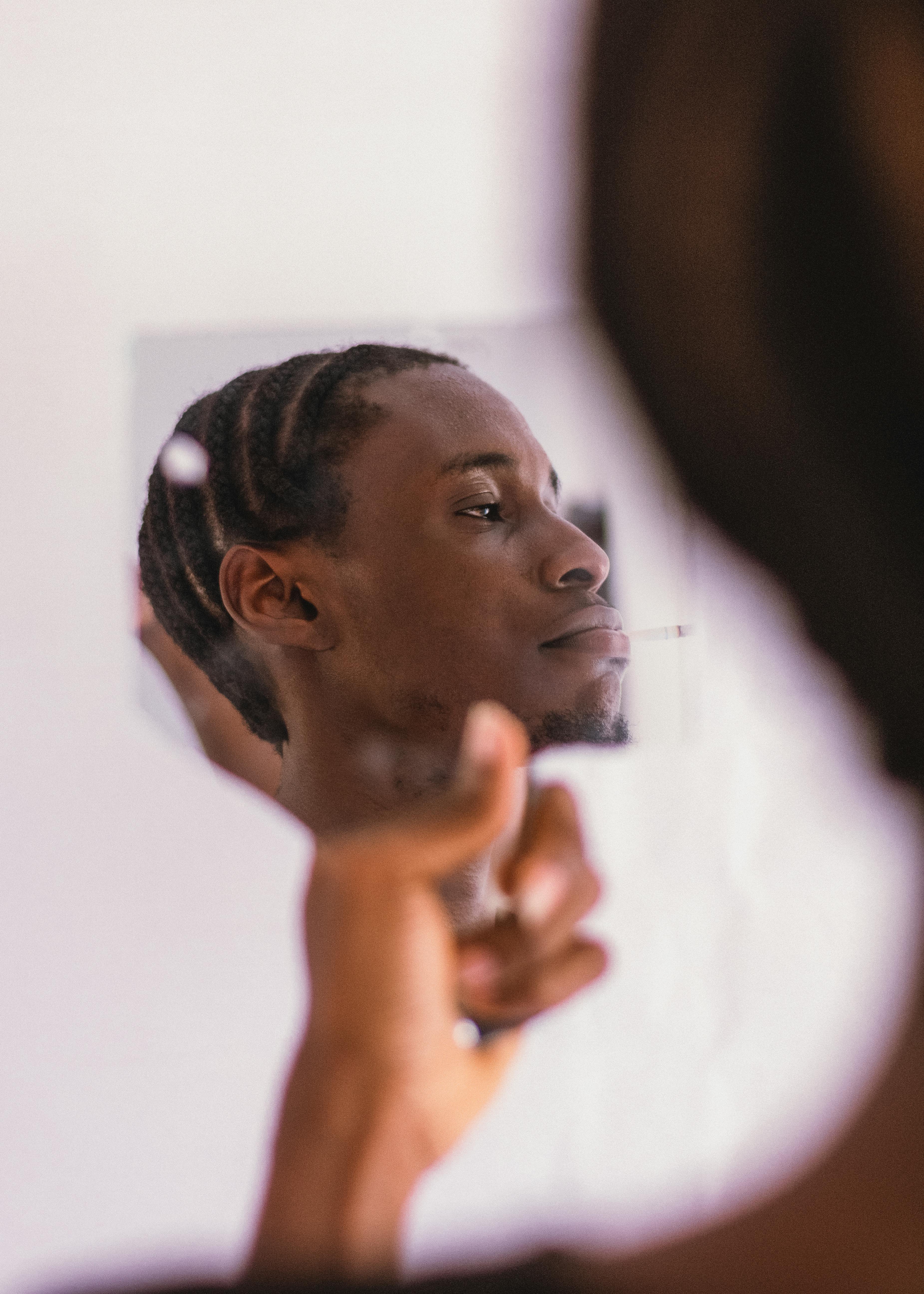 A man with braided hair looks at his reflection in a handheld mirror indoors.