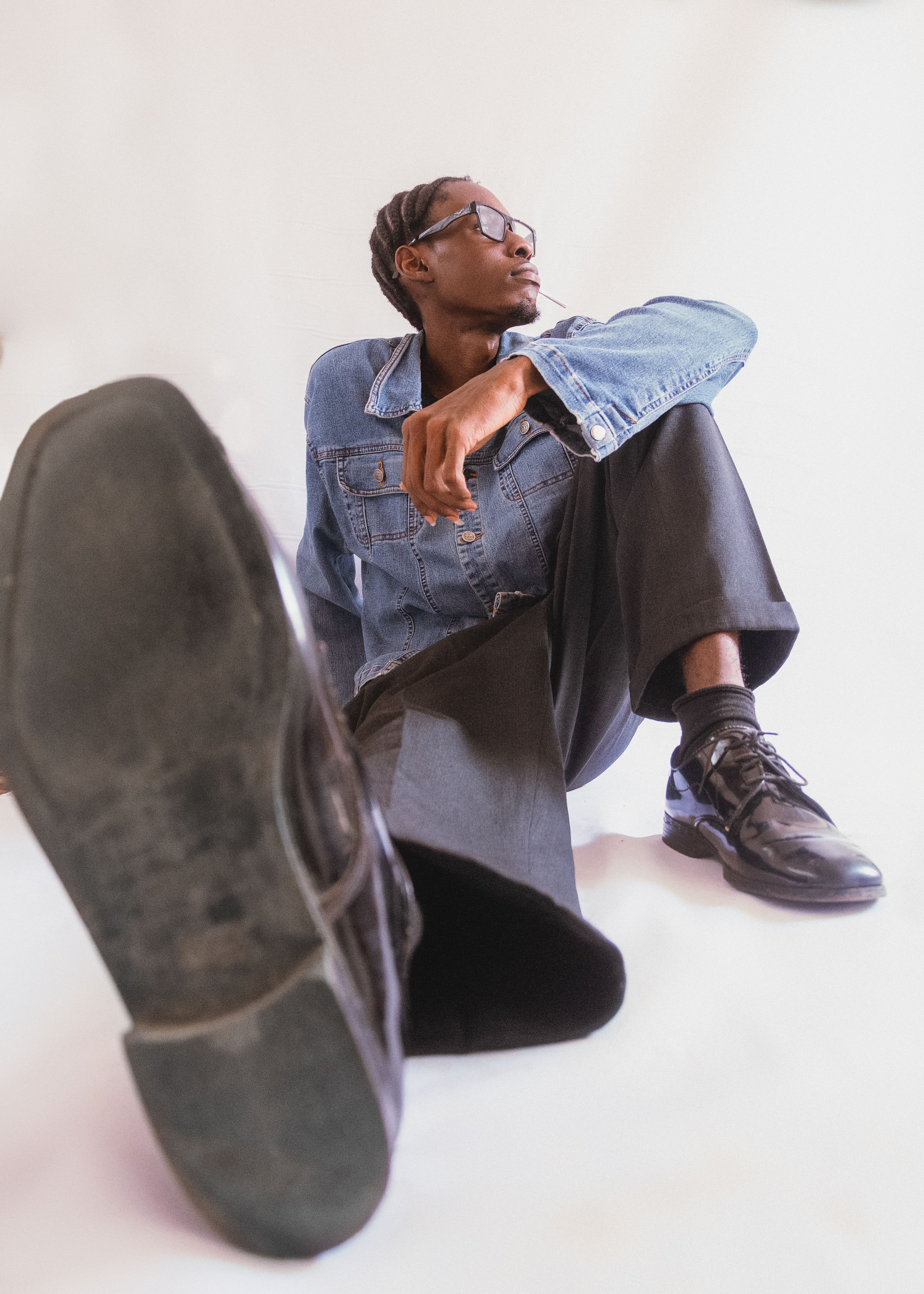 Stylish young man in denim jacket and sunglasses sitting casually against a white background.