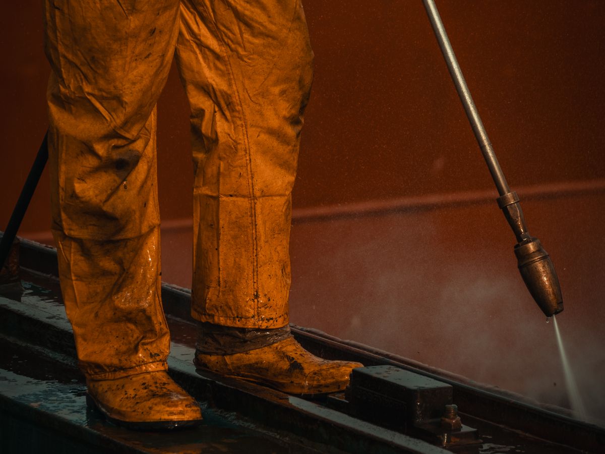 A worker in yellow safety gear uses a high pressure washer on equipment