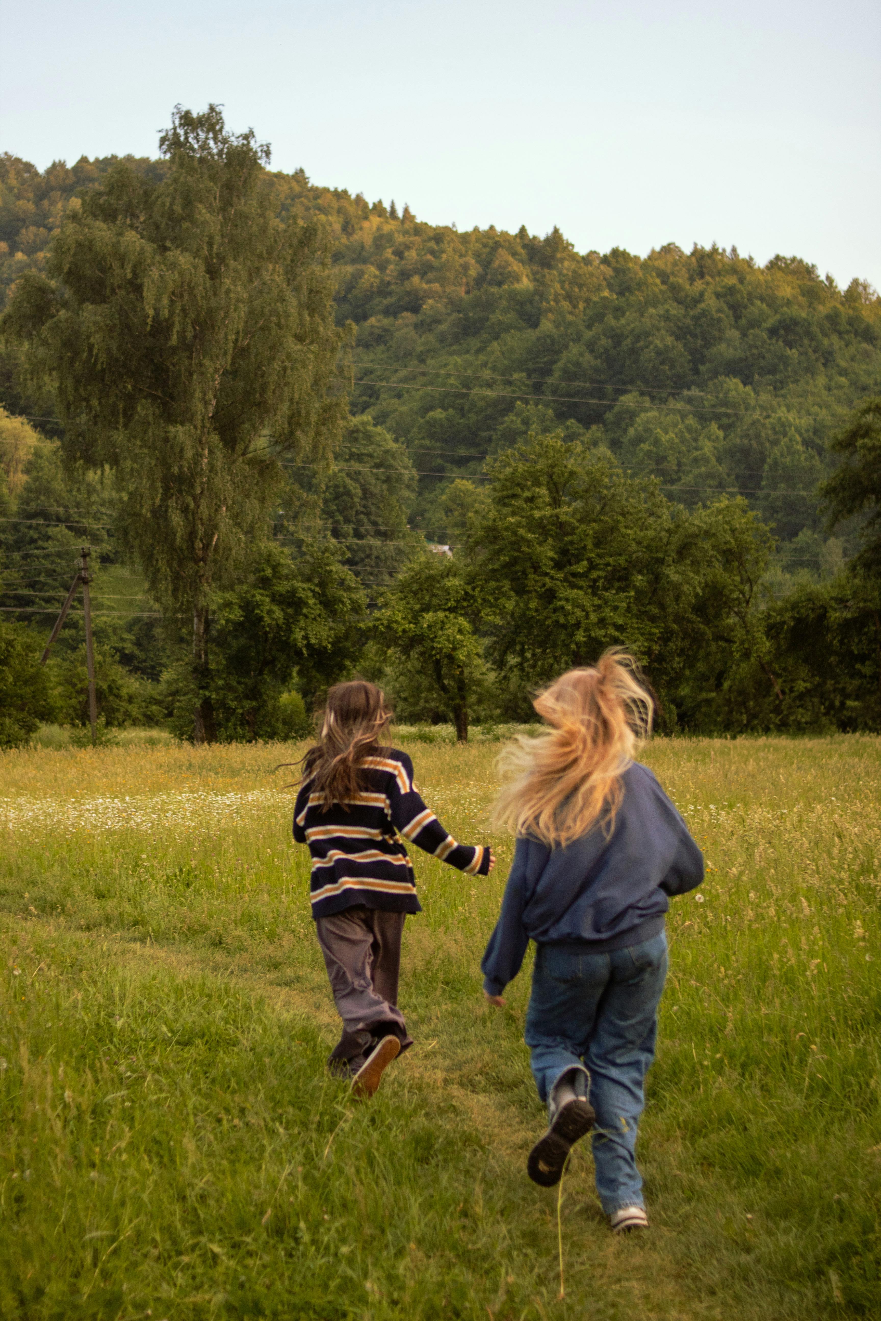 Two children joyfully run through a lush field towards a scenic forest landscape.