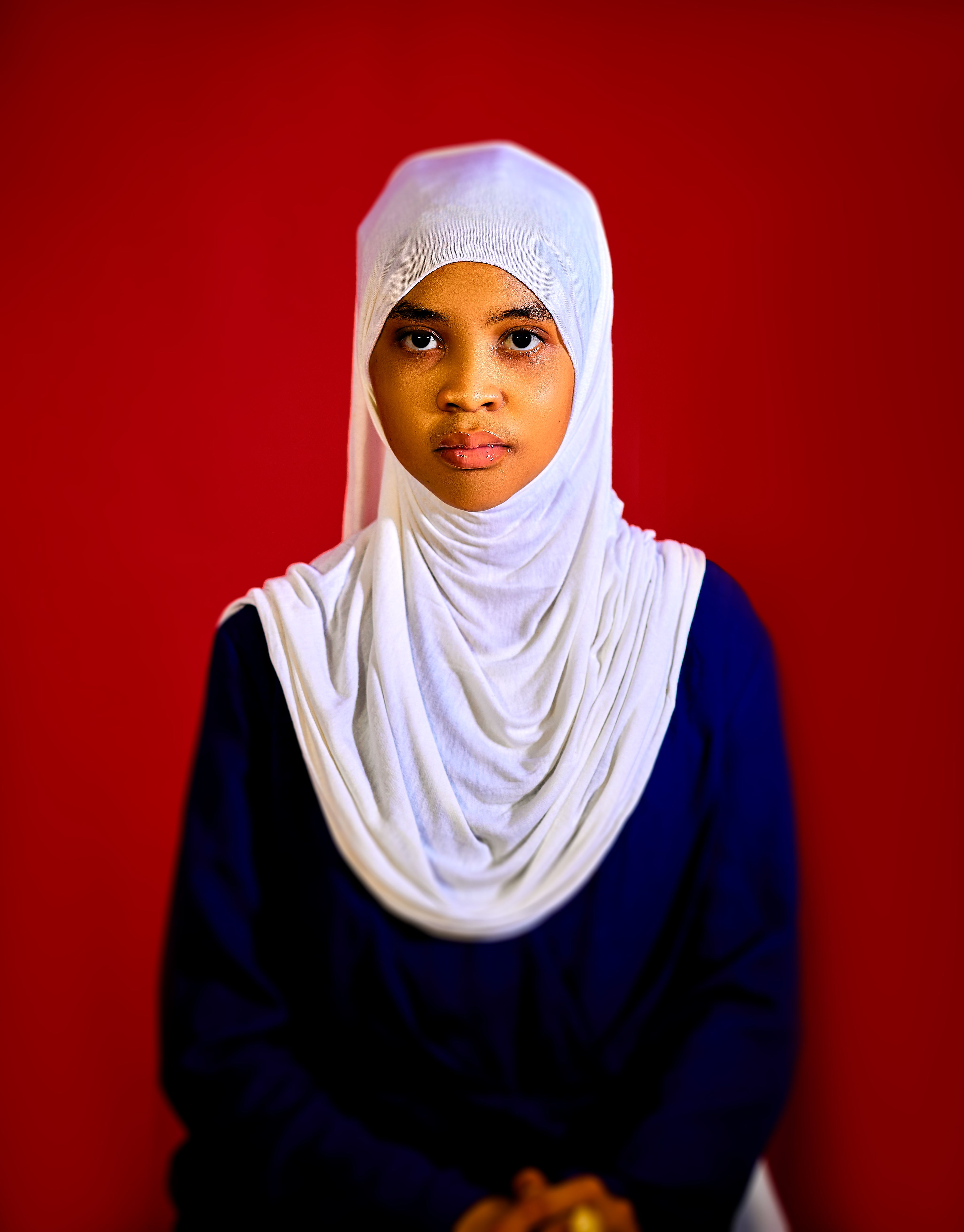 Elegant portrait of a woman wearing a white hijab and navy dress, with a vibrant red background.