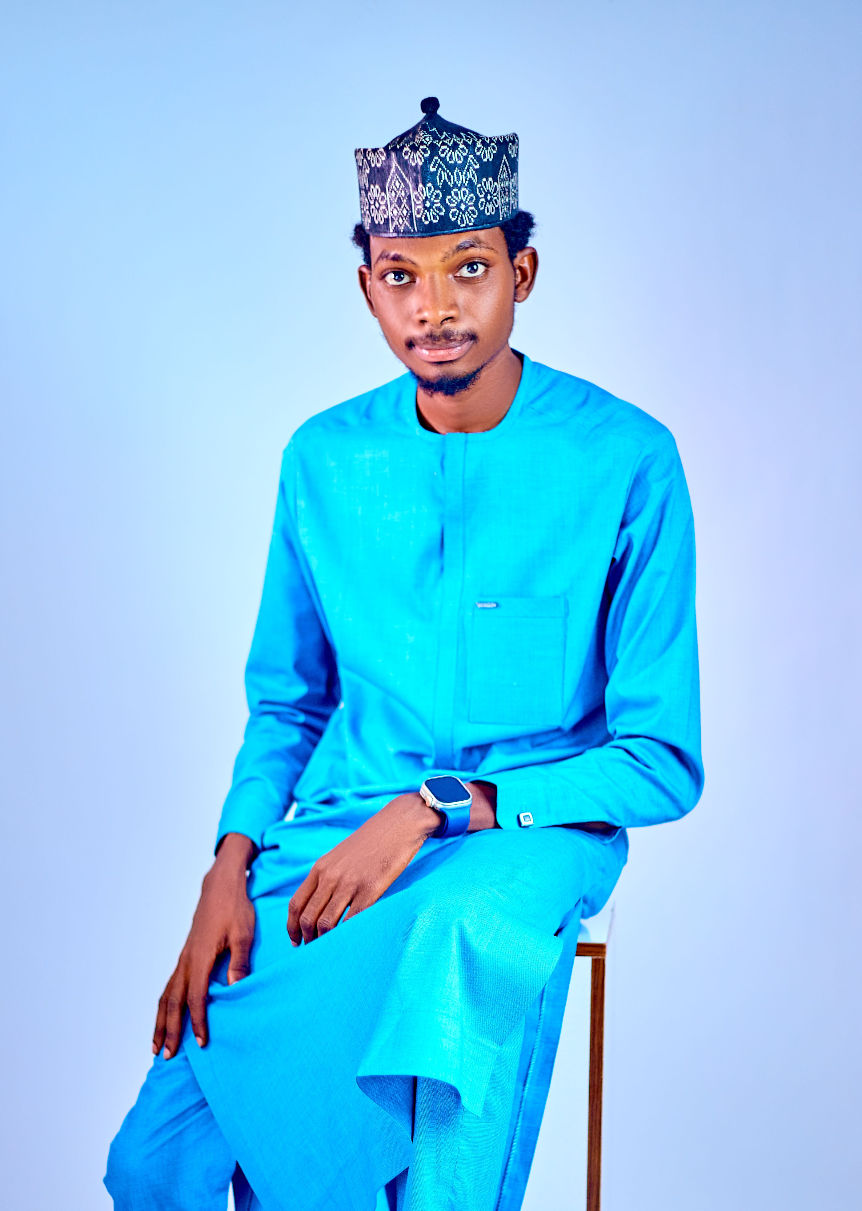 Man in traditional blue outfit seated against a light blue backdrop.