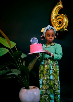 Young girl in traditional dress holding a birthday cake with a balloon in the background.