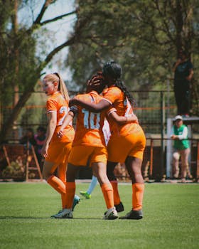 A women's soccer team in orange jerseys celebrates a goal during a match in the bright daytime.