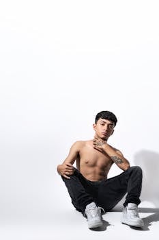 Shirtless young man sitting in casual pose against a bright white backdrop, highlighting modern fashion style.