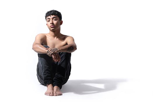 Shirtless young man with tattoos sitting in a thoughtful pose on a white background.