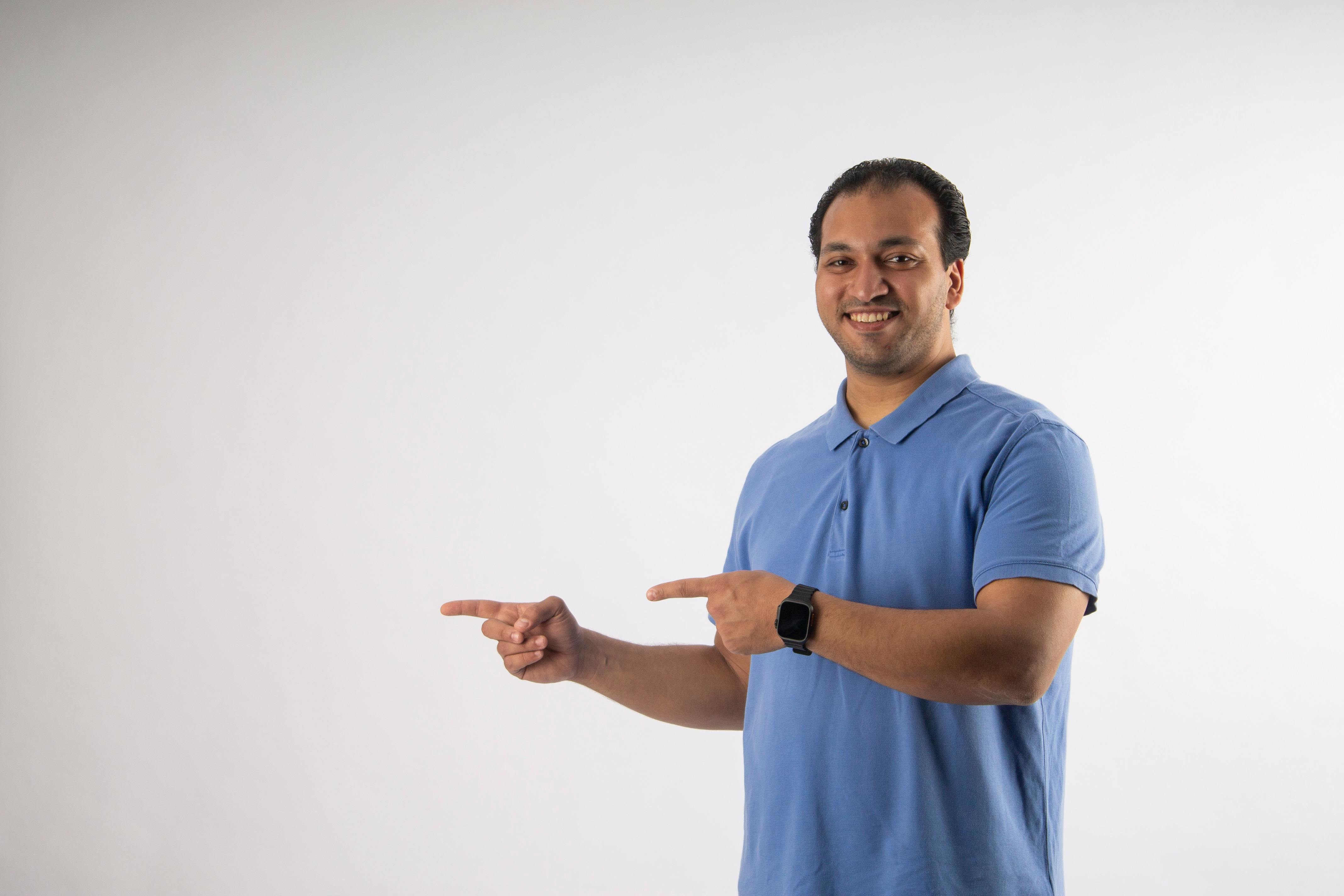 Ilmainen Hymyilevä mies sinisessä poolopaidassa osoittaa sivuttain tyhjällä taustalla. Kuvapankkikuva